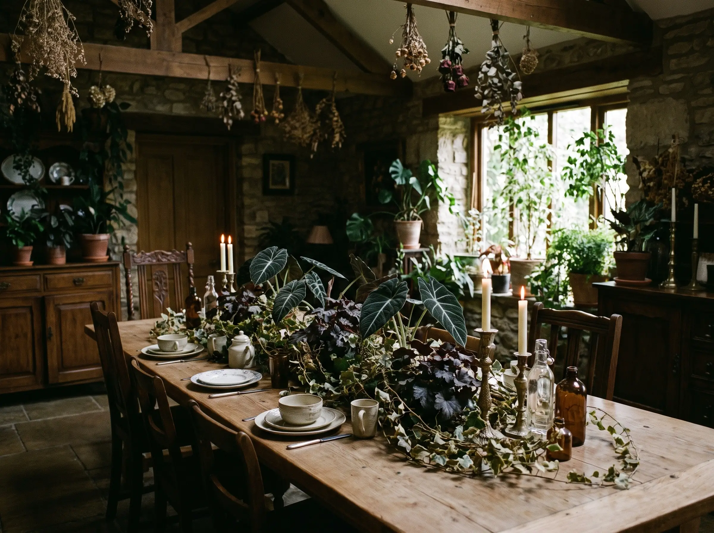 A creeping botanical centerpiece of Black Velvet Alocasia, dark Heuchera, and dried ivy vines runs down a wooden dining table between candle bases.