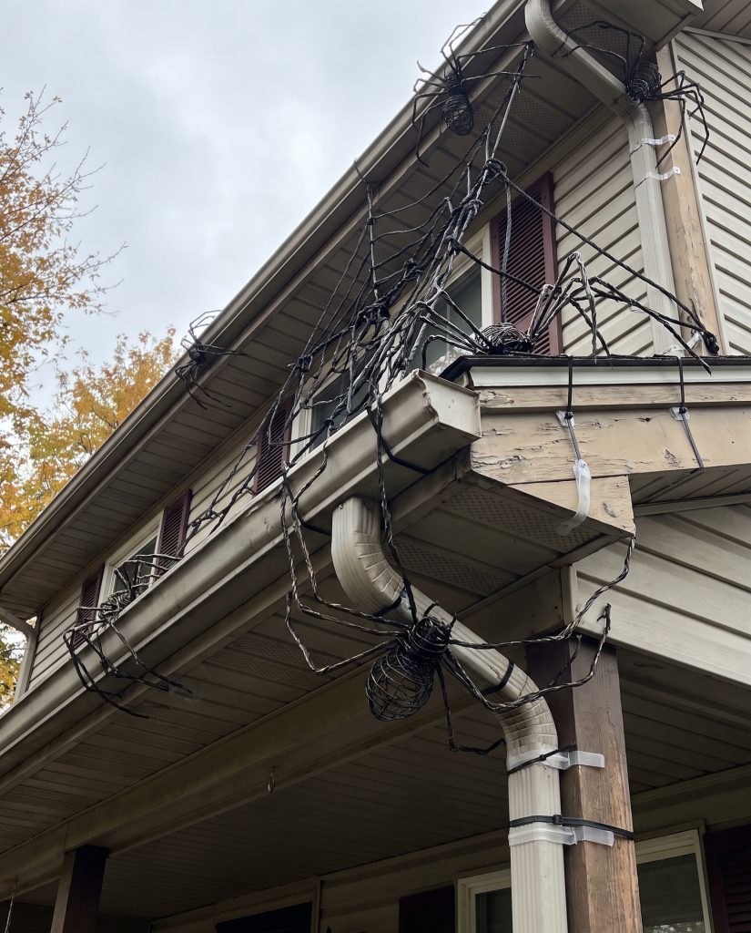 Massive five-foot posable wire spiders cling to the roof eaves and gutter lines of a suburban home for 2026 outdoor Halloween decorating.