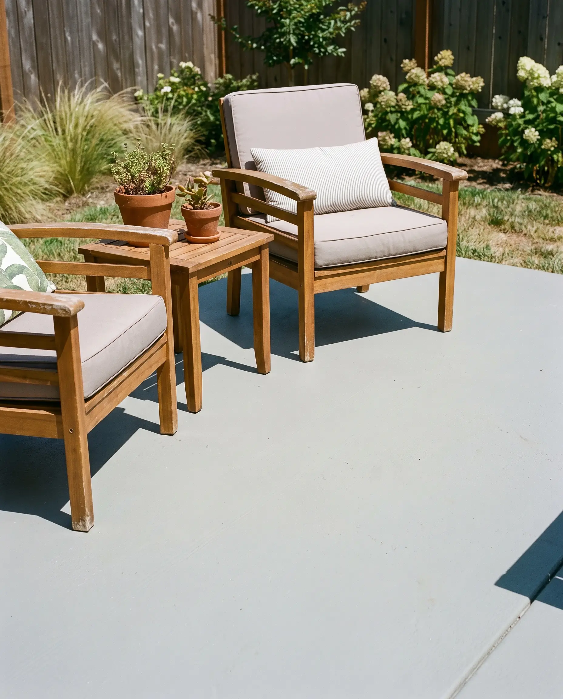 Warm teak wood outdoor chairs and an acacia wood side table rest on a concrete slab coated with the best Behr patio paint colors.