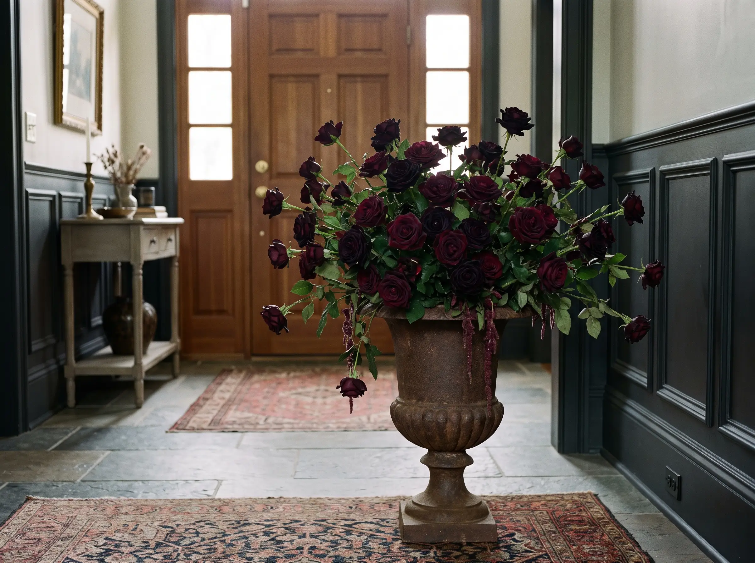 Velvety Black Baccara roses fill a heavy vintage cast iron urn on a modest console table for elegant 2026 halloween decor against charcoal wainscoting.