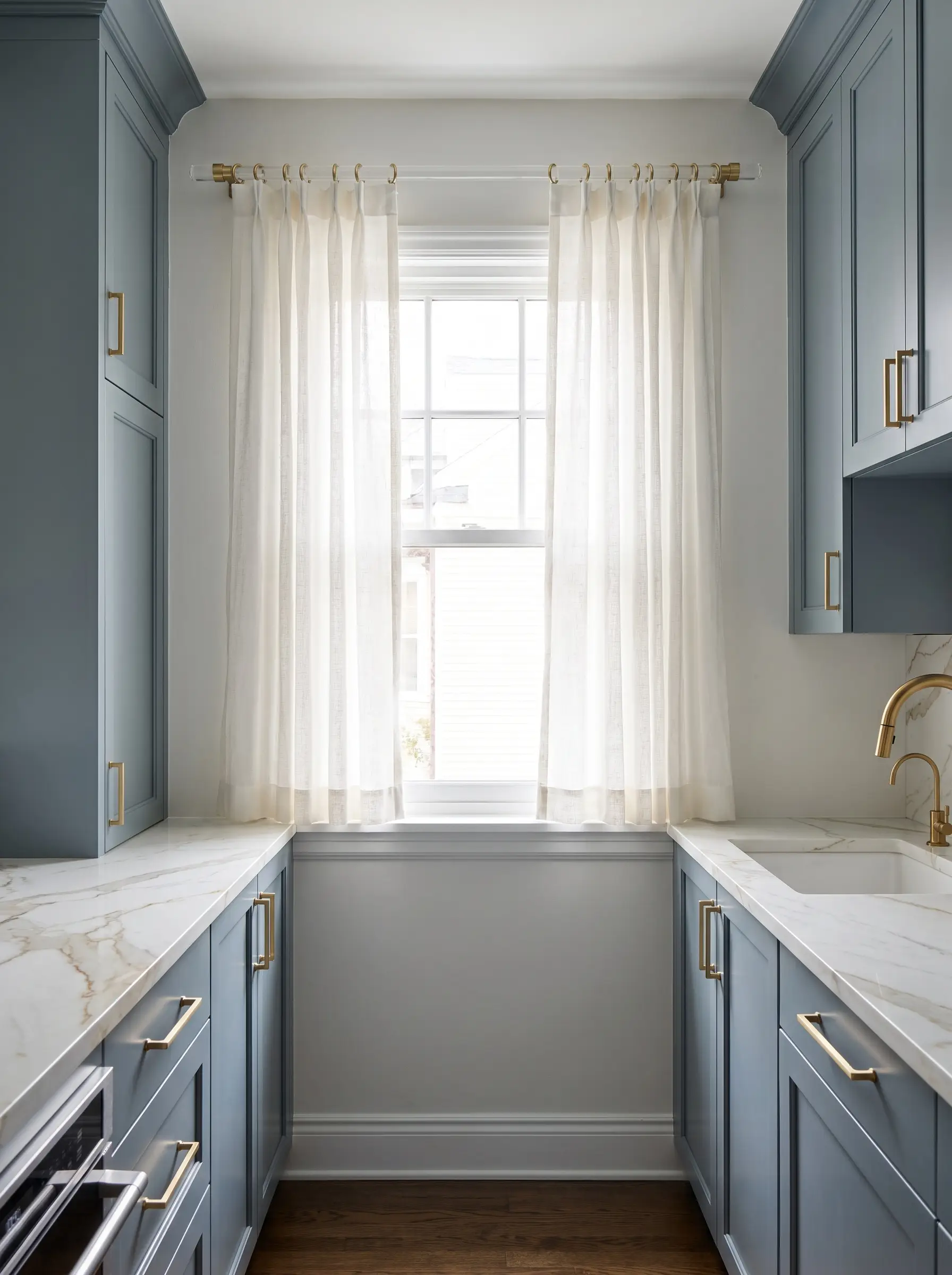 A solid cast acrylic curtain rod with unlacquered brass end-caps supports lightweight linen curtains above a marble-look quartz counter in a galley kitchen.