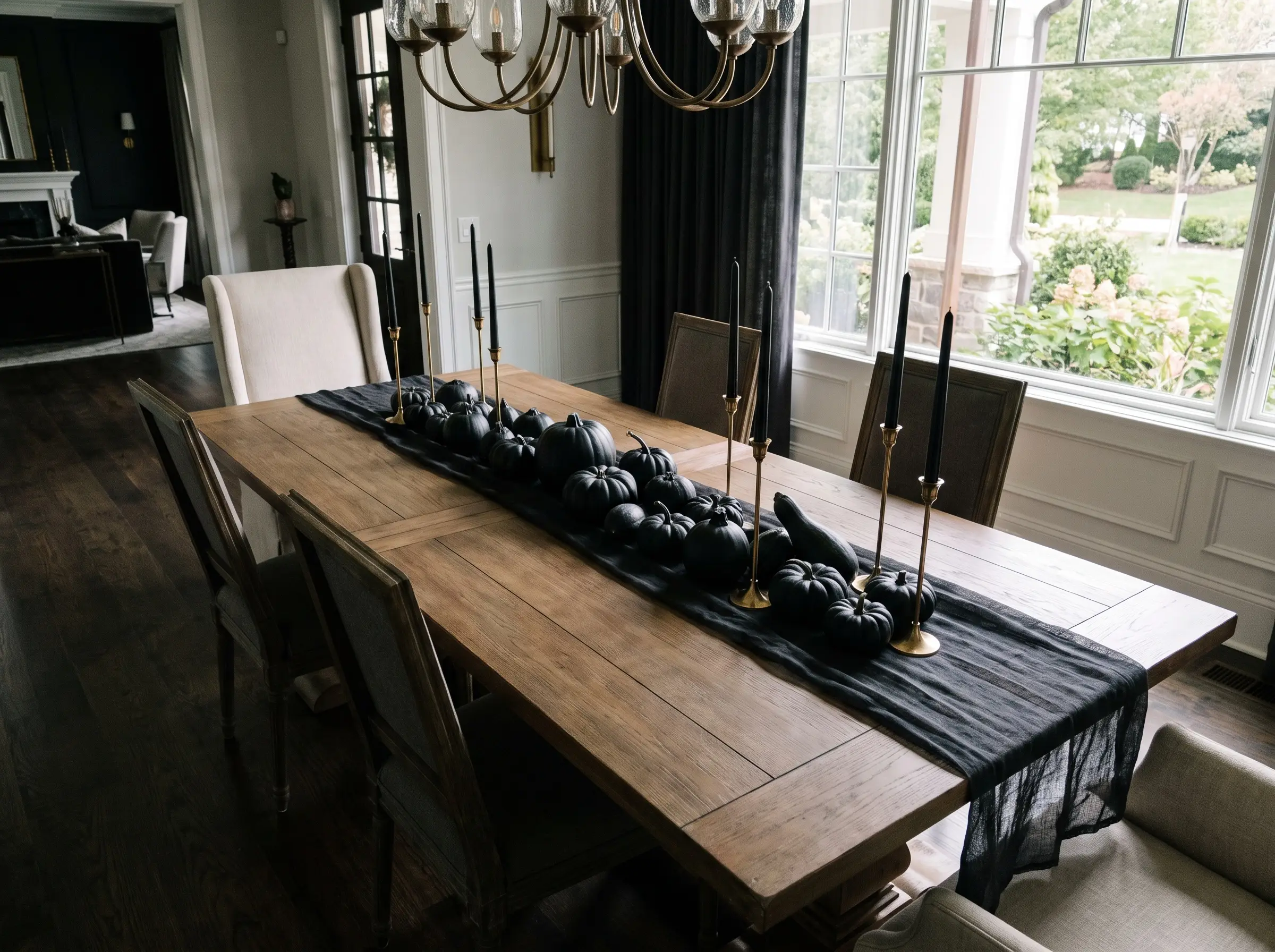 Varying sizes of matte black mini-pumpkins and tall slender brass candlesticks arrange dramatically over a dark charcoal sheer linen runner on a dining table.