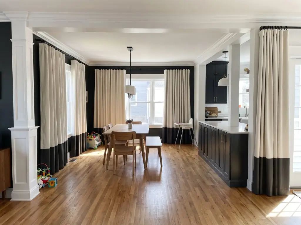 Heavy cotton canvas panels color-blocked in Alabaster and Greenblack hang from matte black rings on a sleek French return rod over warm wood flooring.