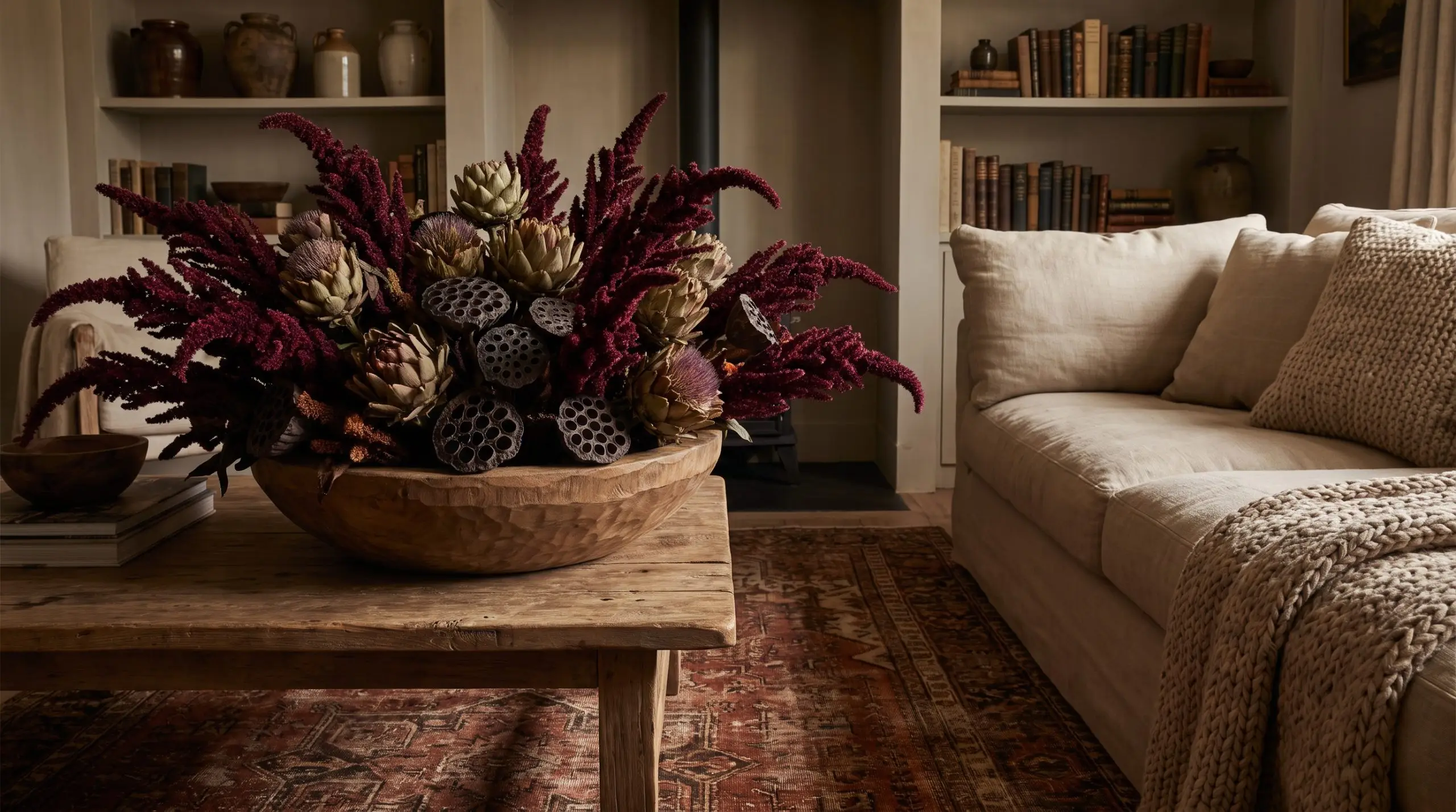 Deep-burgundy amaranth, dried artichokes, and dried lotus pods sit in a wide wooden bowl on a standard coffee table near a linen sofa.