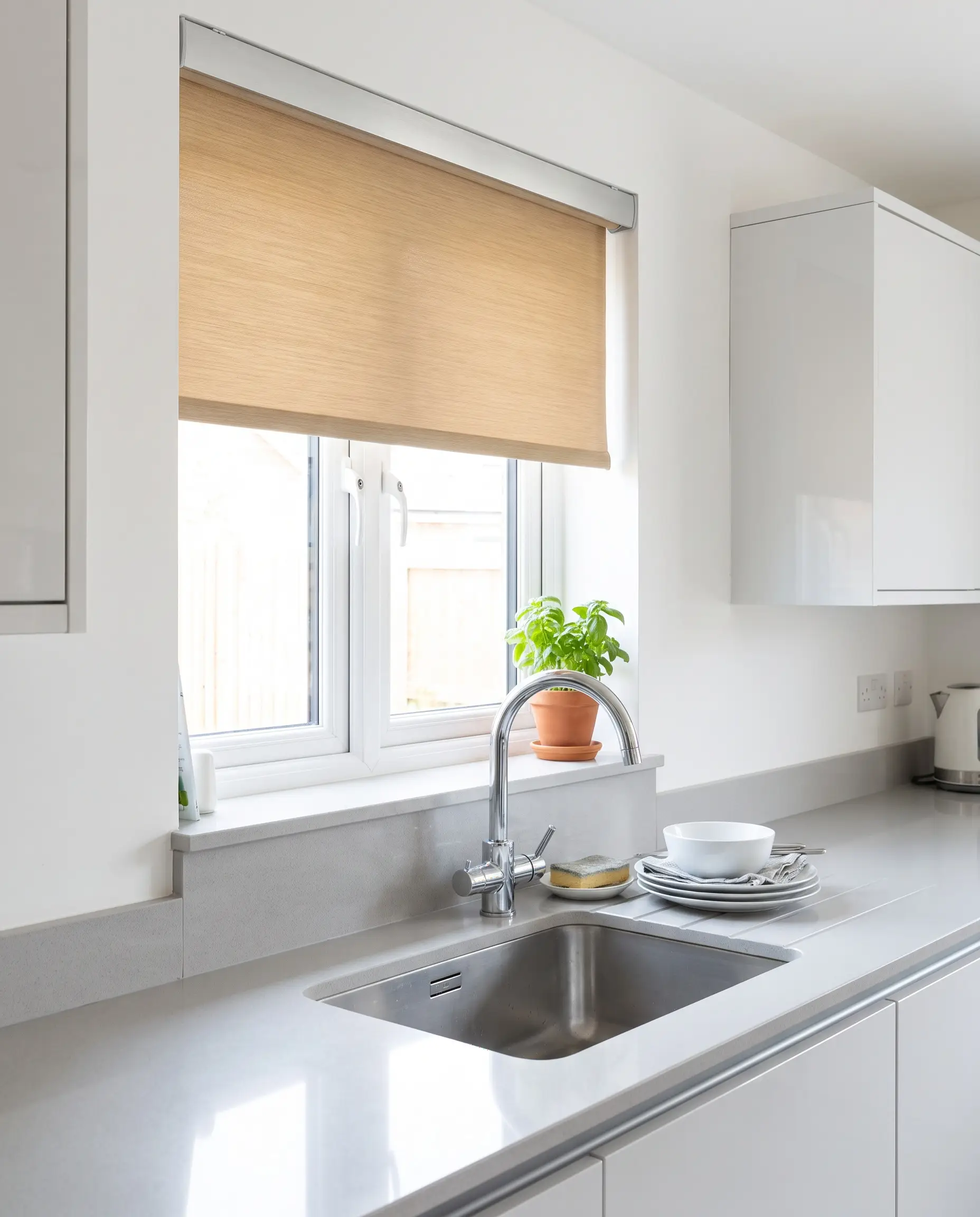 A slim-profile faux wood roller blind retracts into a minimal aluminum fascia above a sleek quartz sink countertop and modern chrome gooseneck faucet.