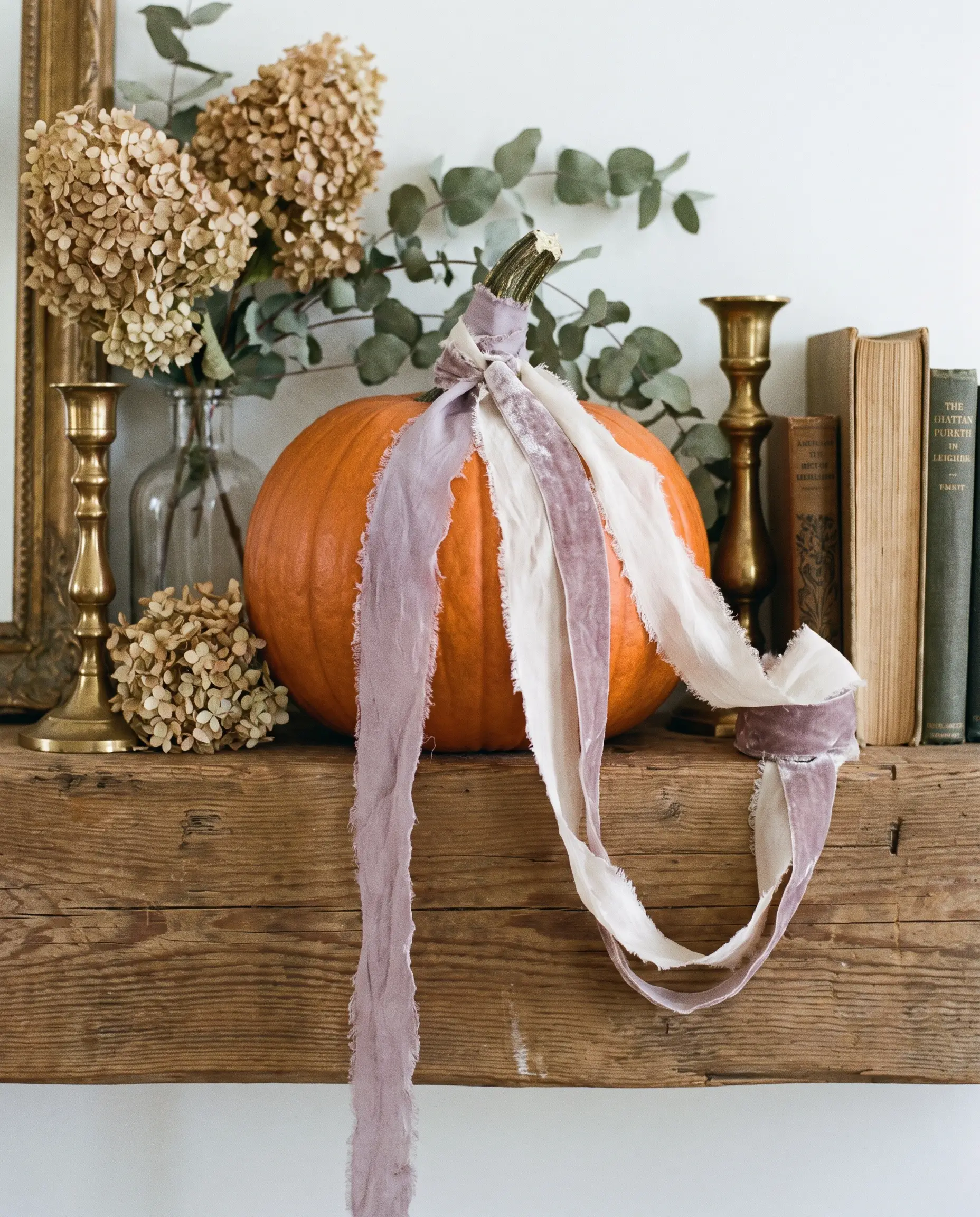 Frayed, hand-torn silk and crushed velvet ribbons in dusty lavender and clotted cream tie elegantly around a pumpkin stem on a standard wooden mantel.