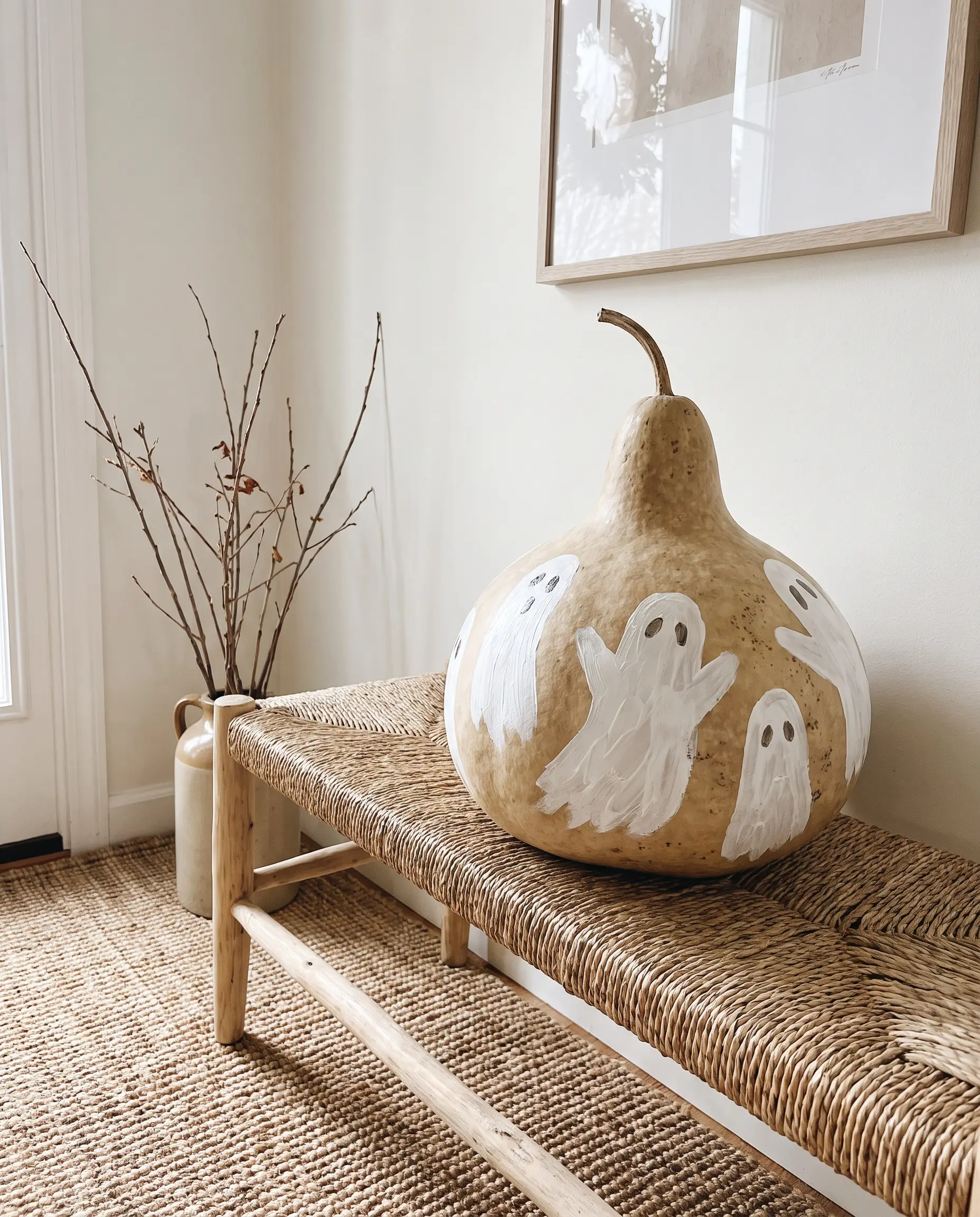 Sweeping minimalist ghost silhouettes painted in heavy-body white acrylic decorate a raw, natural beige gourd positioned on a standard wooden entryway bench.
