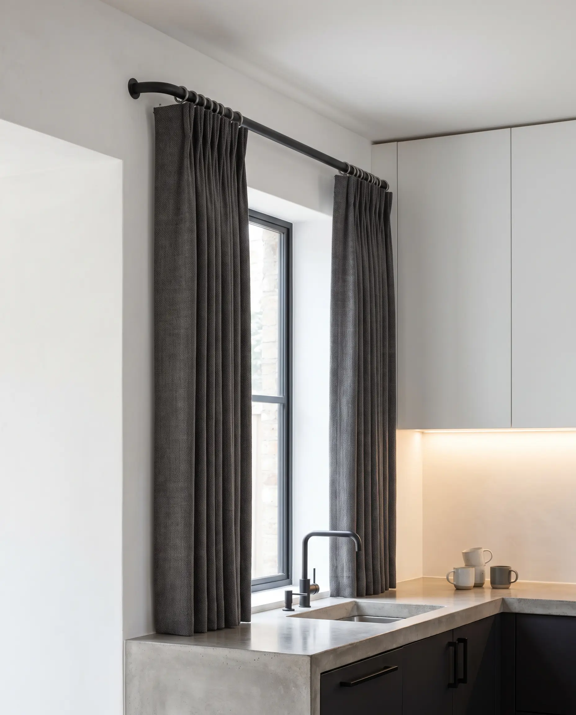 Tailored pinch pleat drapes hang from a powder-coated matte black steel French return rod curved directly into the drywall above a smooth concrete-finish countertop.