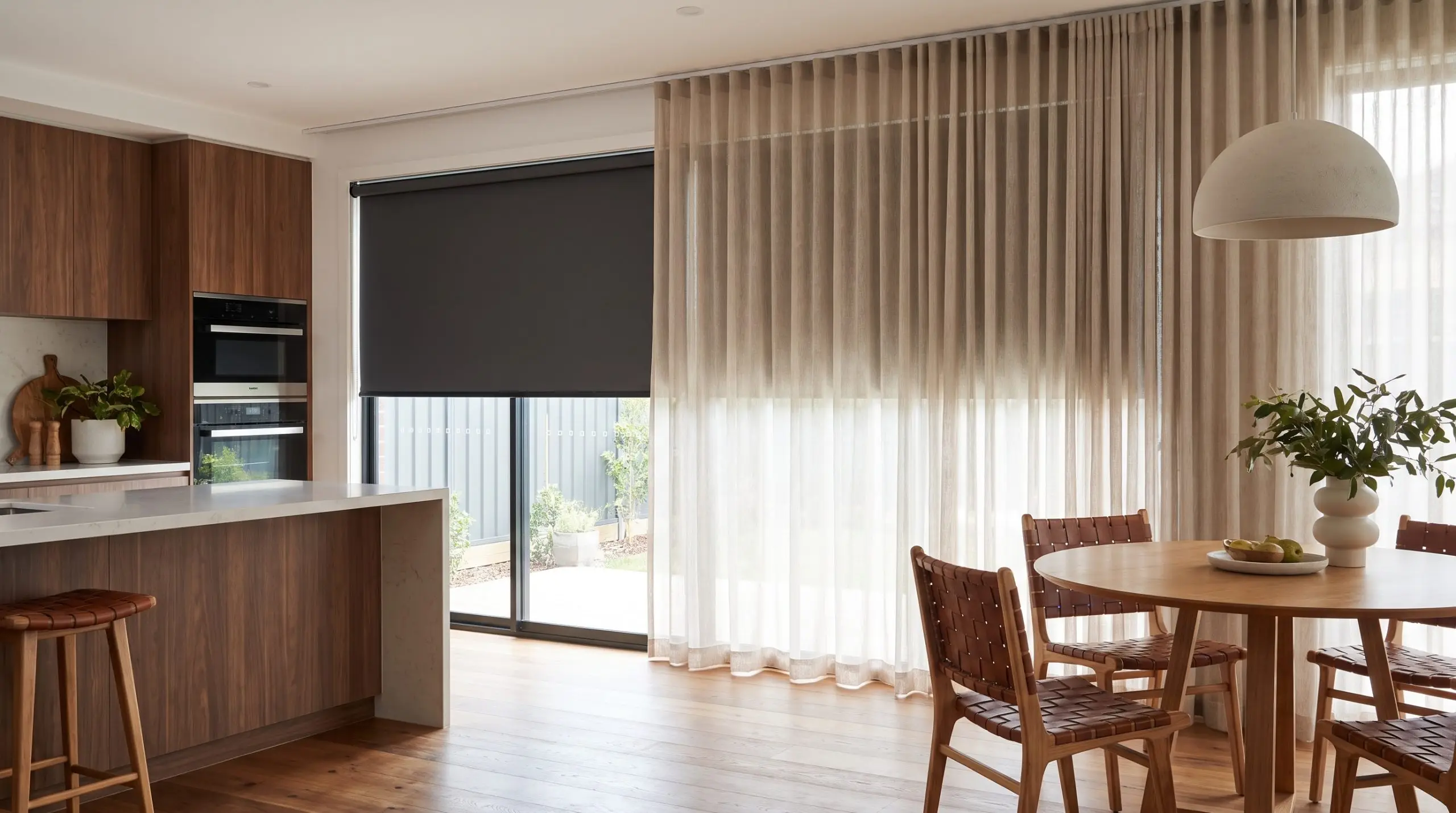 Traverse sheer performance linens cascade over a matte blackout roller blind on a dual-track ceiling mount beside a round dining table and woven chairs.