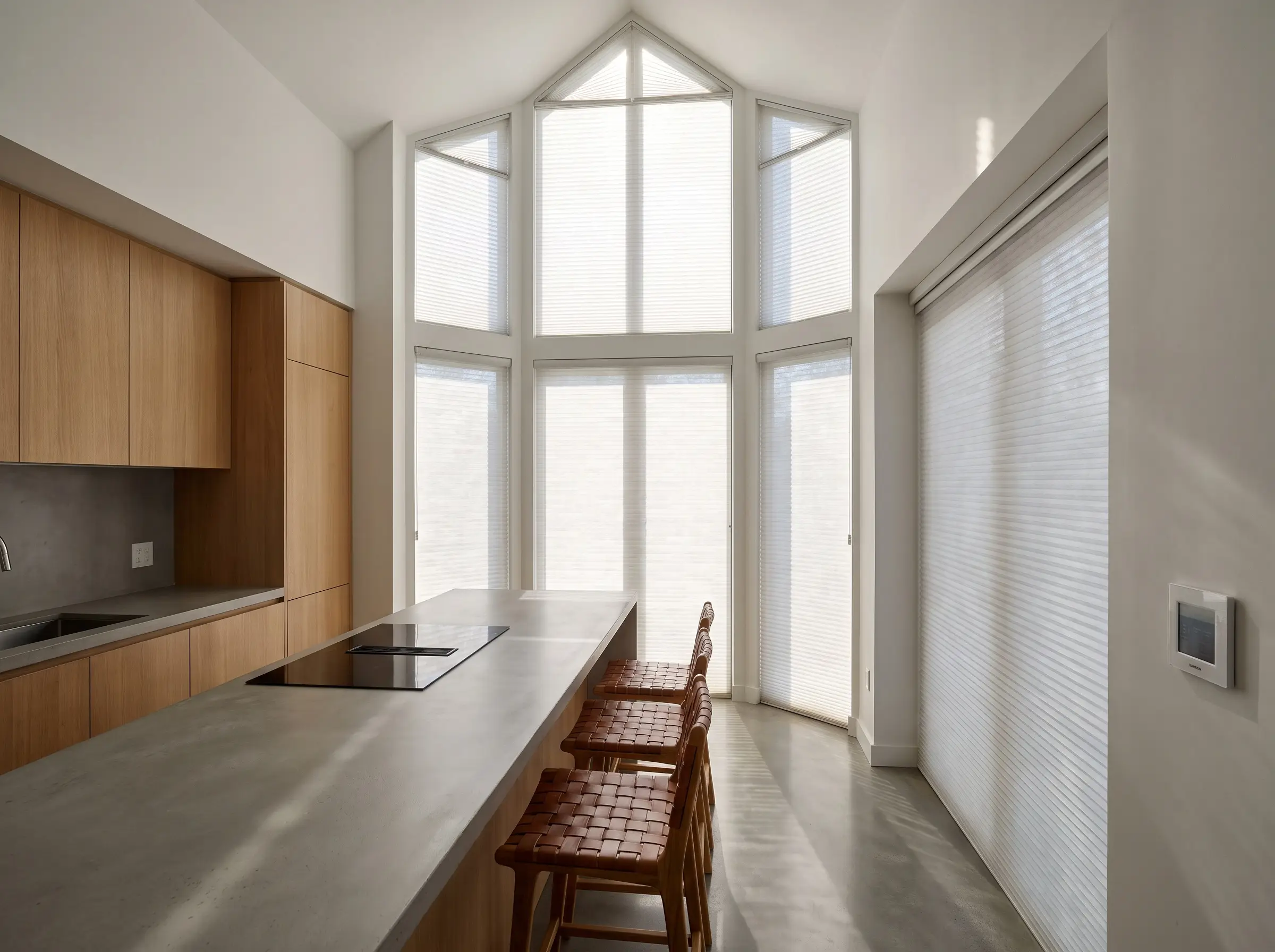 Light filtering motorized cellular shades cover expansive bay windows in a minimalist space featuring handleless cabinetry and a concrete-look kitchen island.