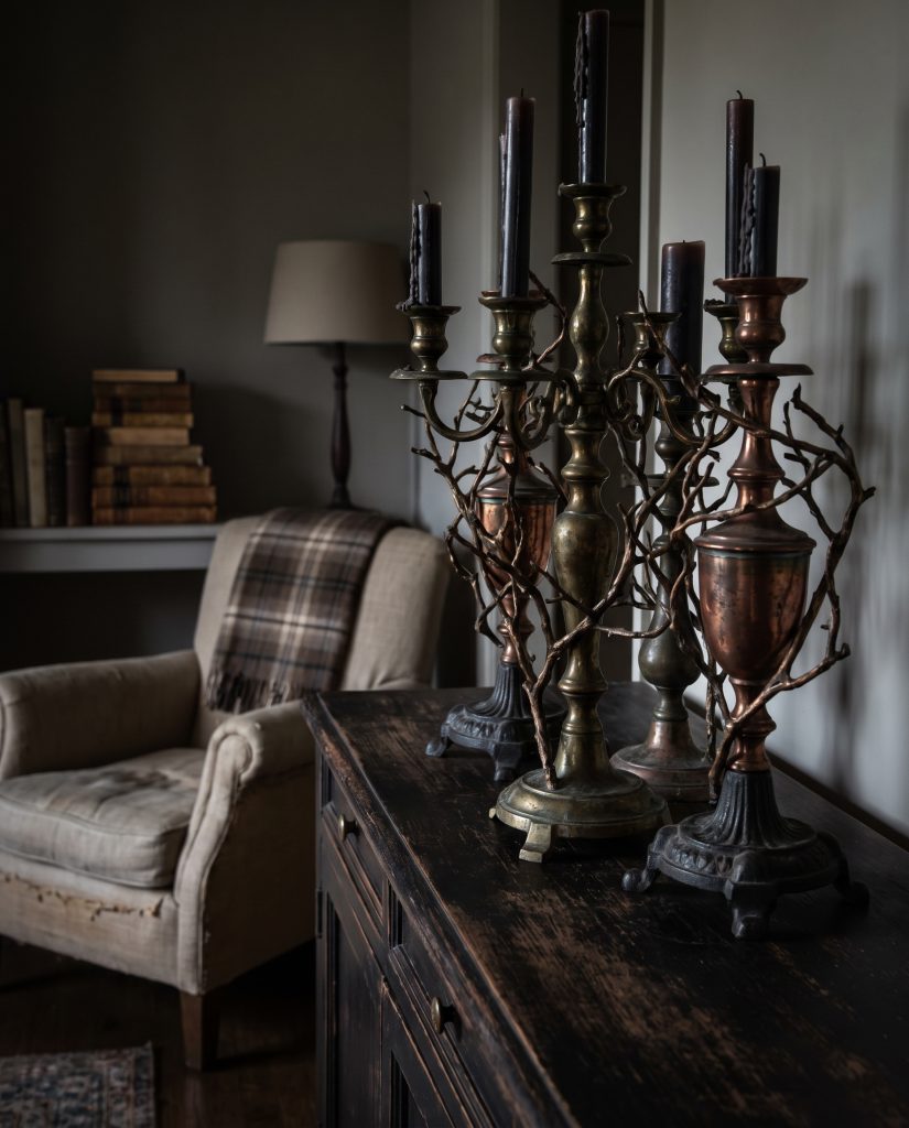 Three heavy patinated brass candelabras rest on a dark wood credenza next to a linen armchair, creating elegant 2026 halloween decor.