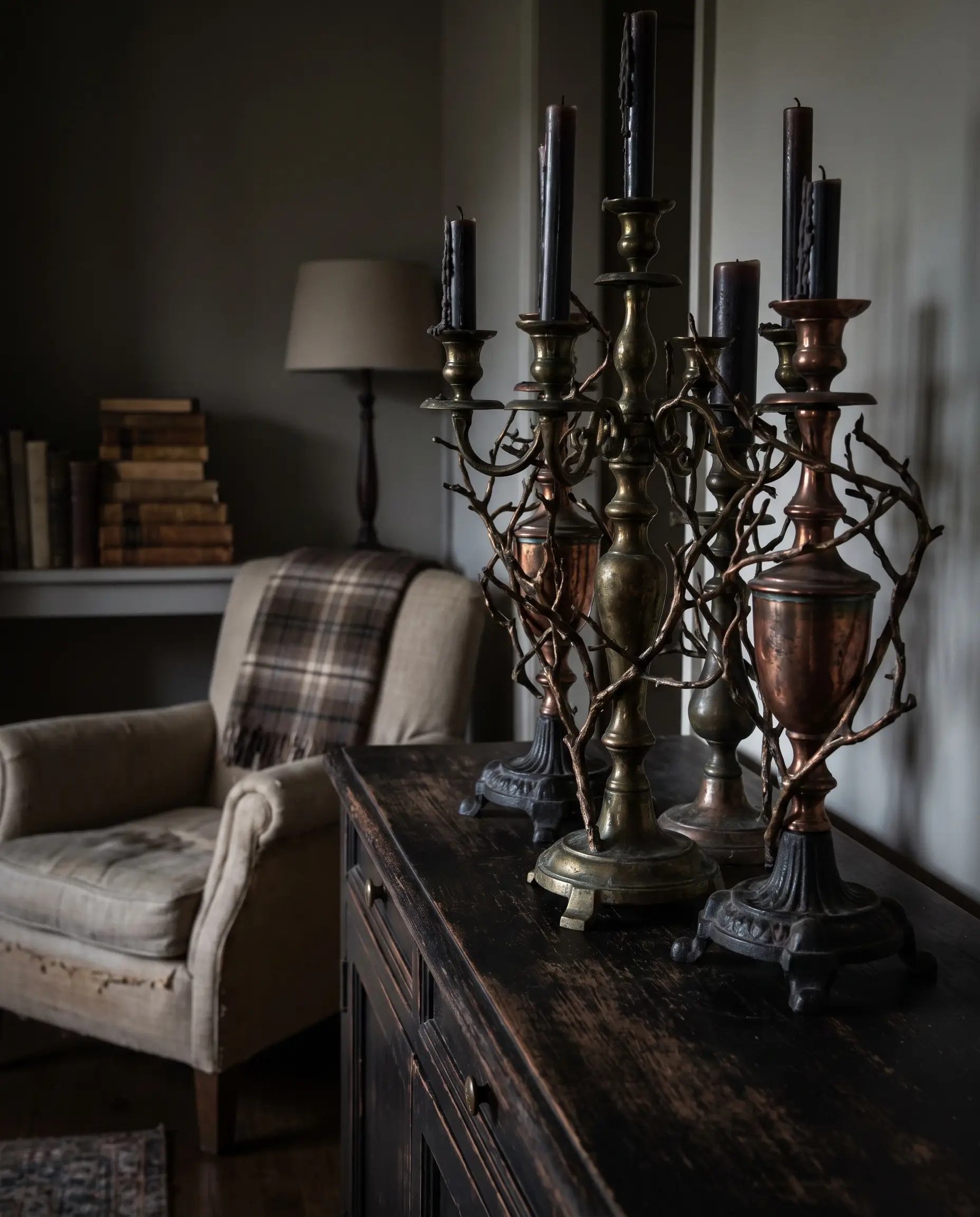 Three heavy patinated brass candelabras rest on a dark wood credenza next to a linen armchair, creating elegant 2026 halloween decor.
