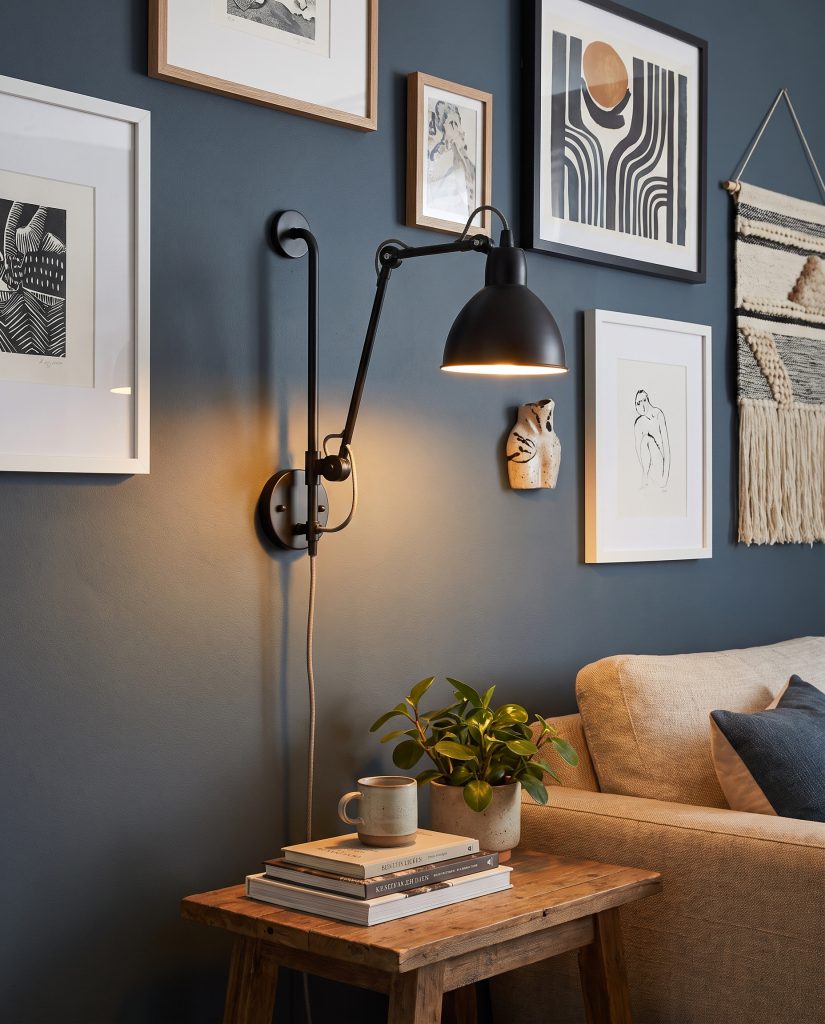 A matte black articulating plug-in sconce casts a warm glow among framed prints on a deep blue gallery wall above a wooden side table.