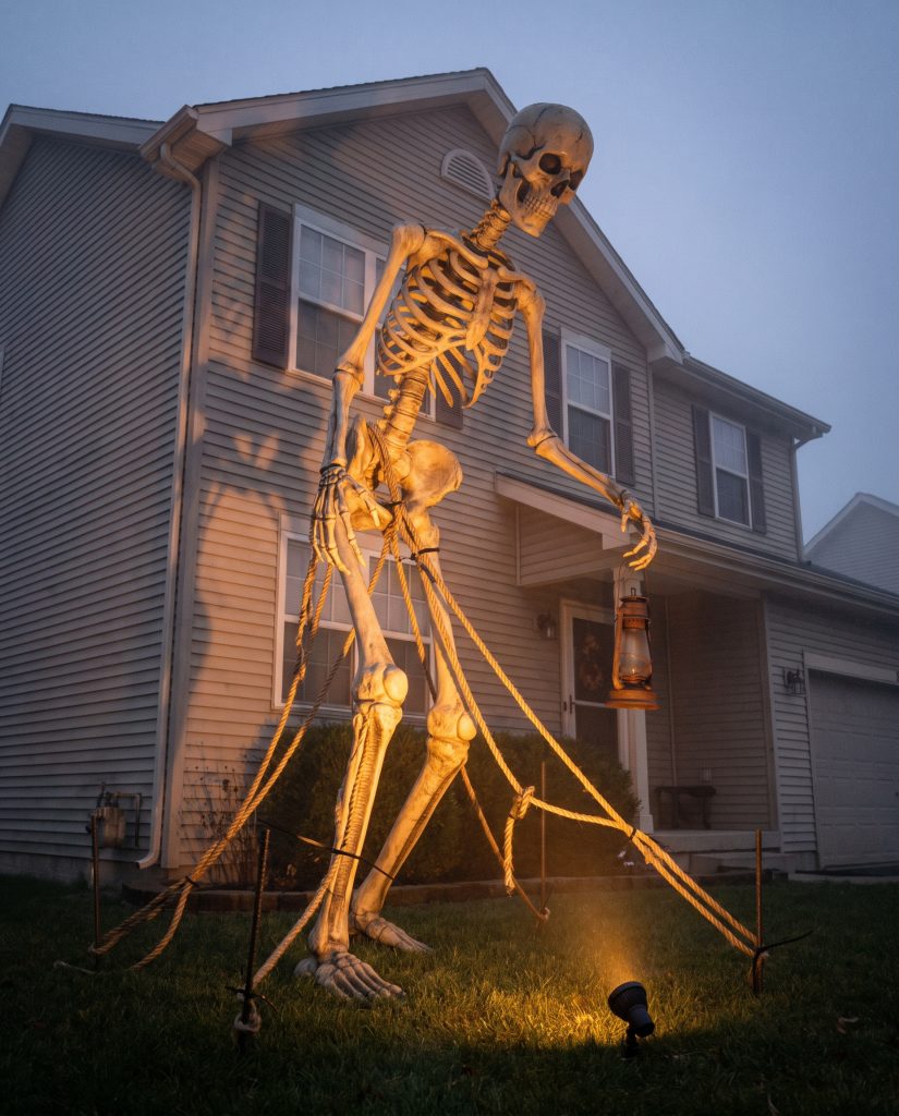 A twelve-foot posable skeleton tangled in thick manila rope holds a heavy iron lantern on a suburban home grass lawn.