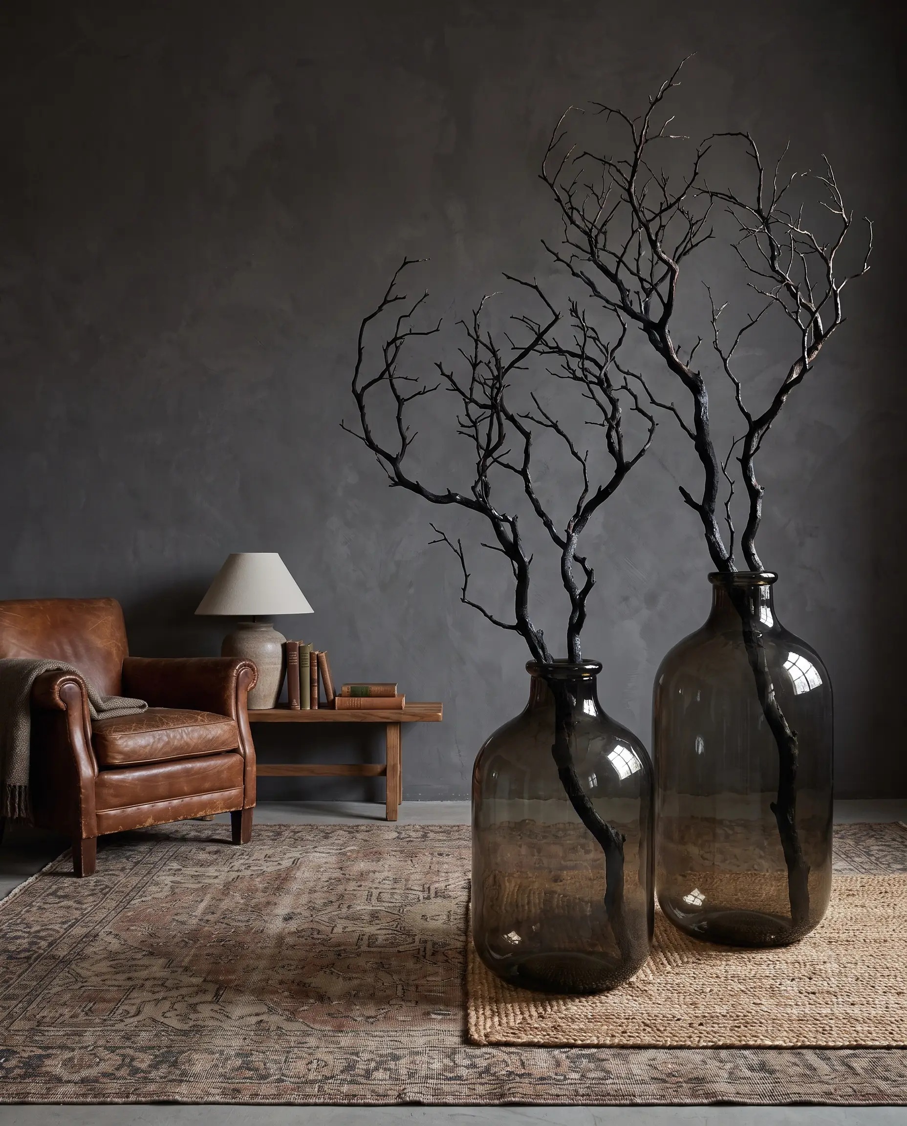 Oversized heavy smoked glass floor vases hold stark, charred manzanita branches against a moody wall next to a vintage armchair and subtle rug.