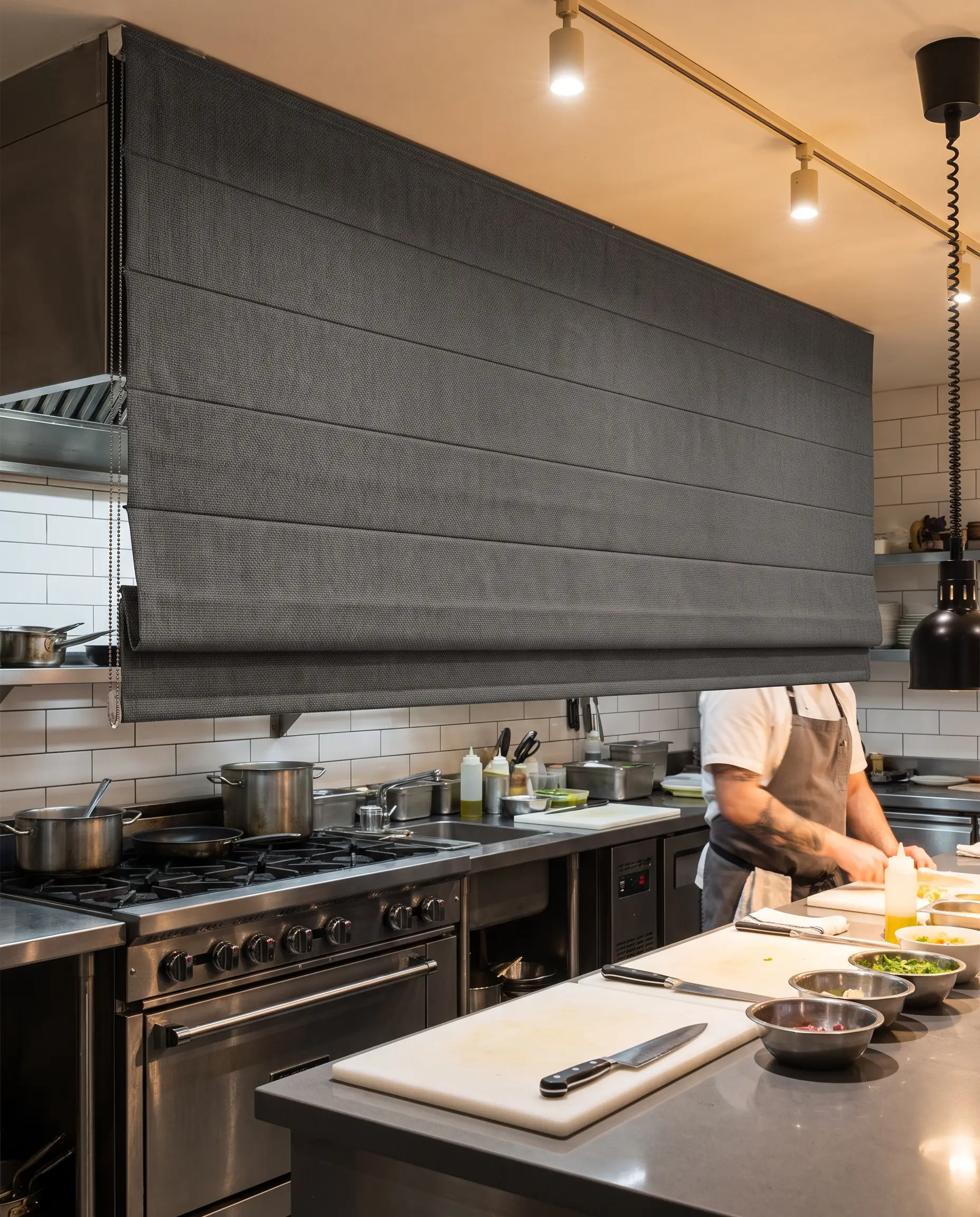 A structured Roman shade crafted from solution-dyed acrylic fabric hangs above a professional-grade stainless steel range, sleek quartz countertops, and minimalist subway tile.