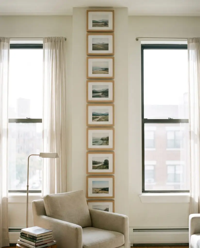 A vertical stack of identical landscape-oriented frames tightly fills a narrow white drywall section between two tall windows near a reading nook.