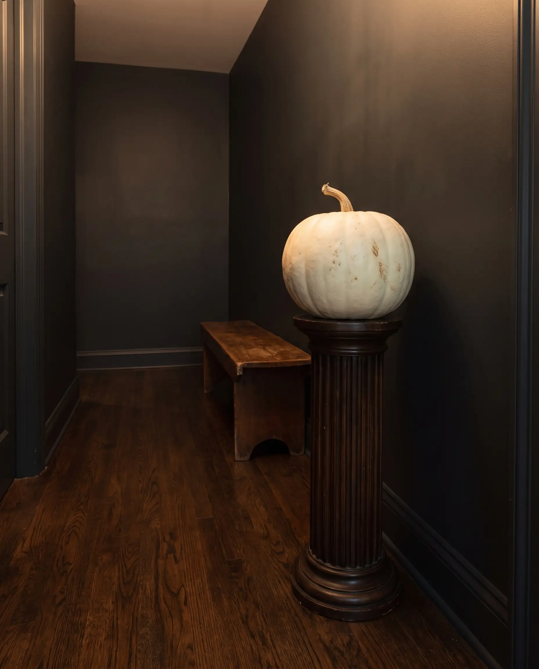 A single white Ghost pumpkin rests on a fluted wood pedestal in a moody hallway corner featuring charcoal-painted walls and sophisticated spooky decor.