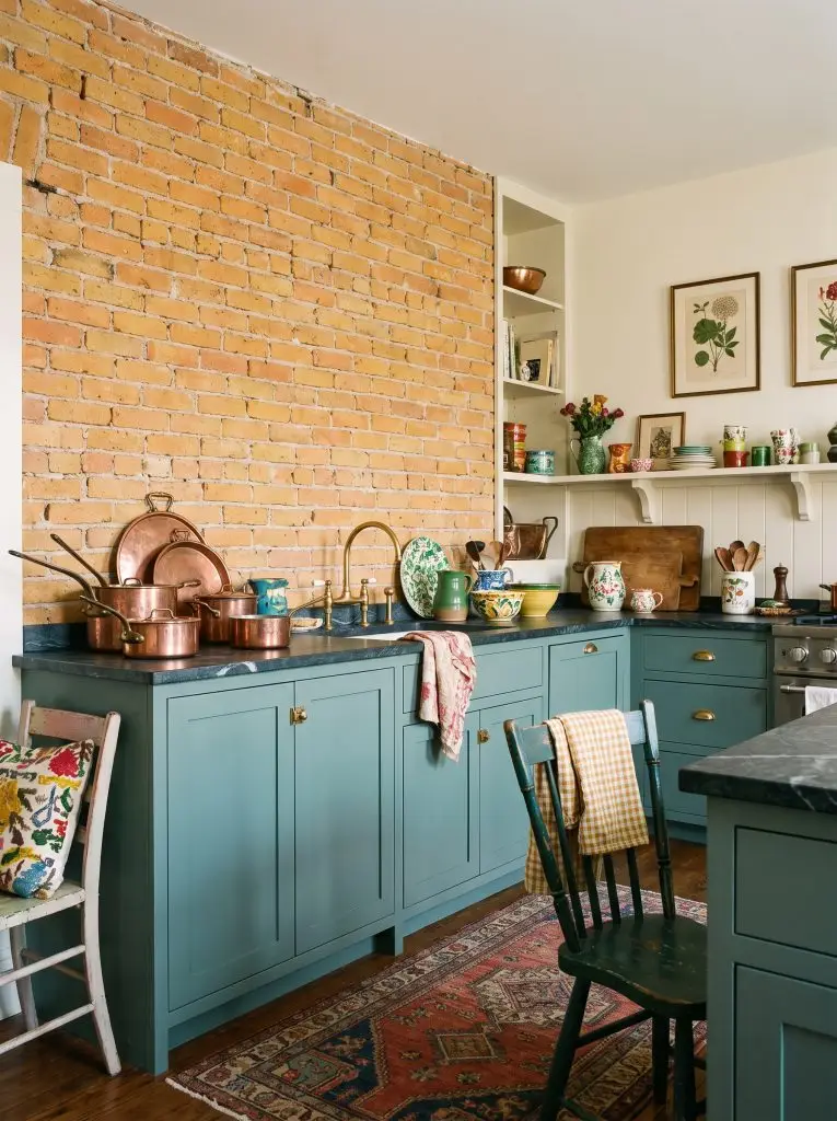 Soapstone counters and unlacquered brass hardware adorn Benjamin Moore Aegean Teal lower kitchen cabinets positioned beneath an exposed orange-yellow brick wall.
