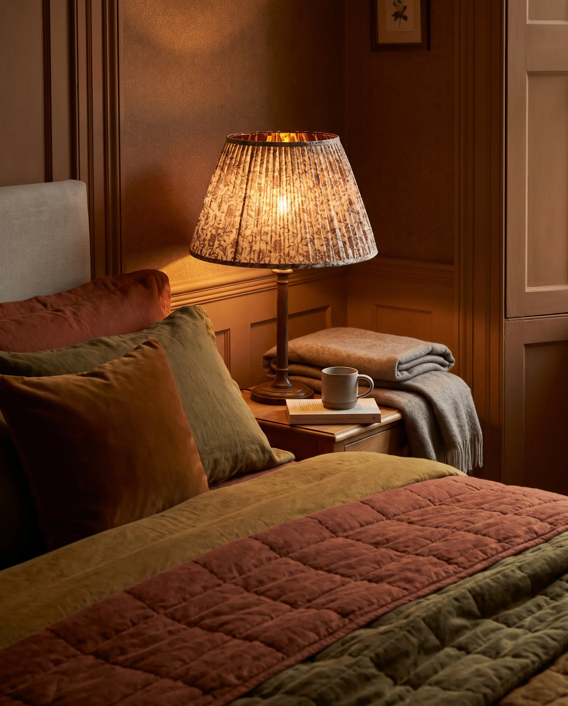A bedside lamp featuring a block-printed cotton pleated shade illuminates a nightstand and heavy linens in a cozy fall bedroom.