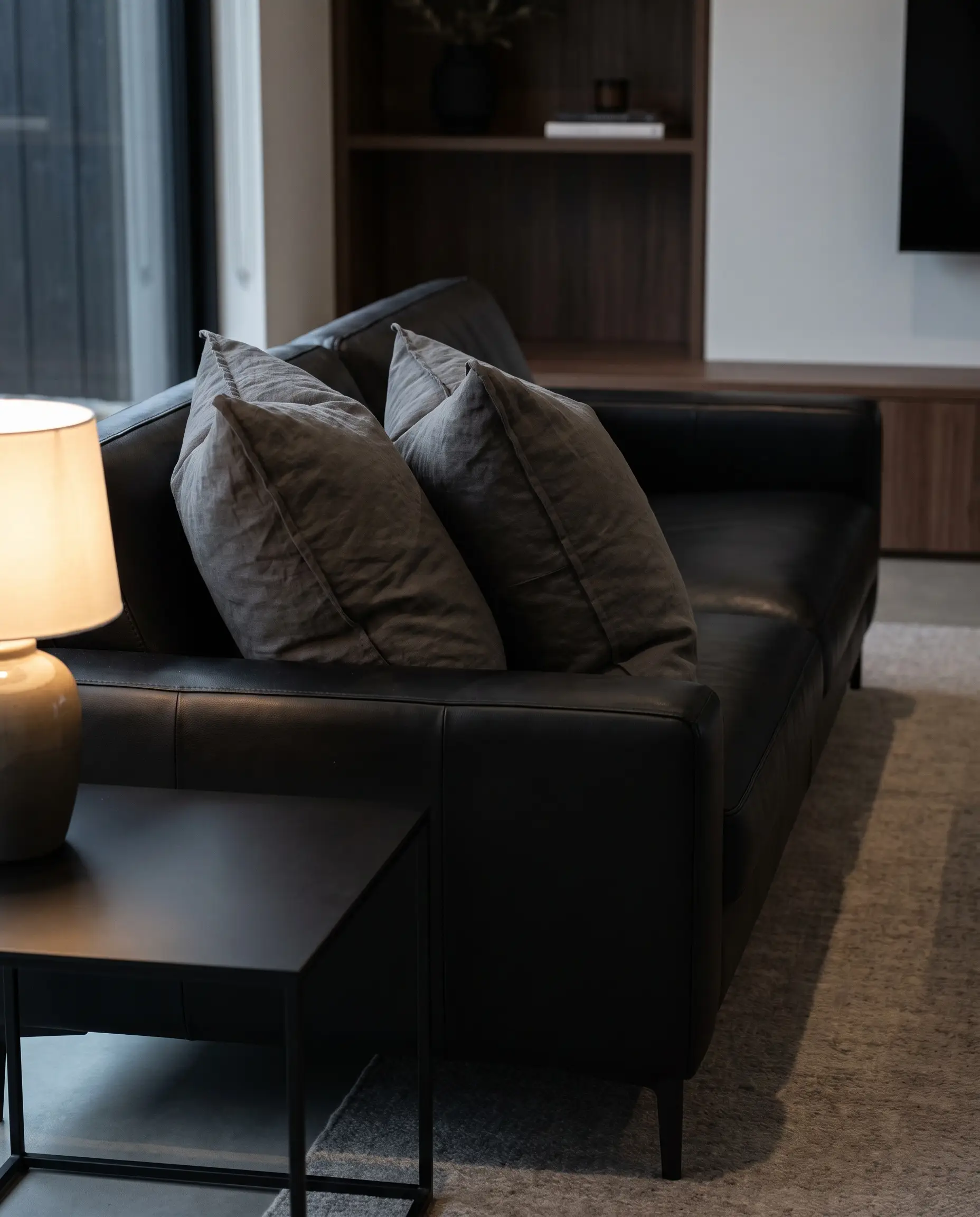 Kendall Charcoal heavyweight Belgian linen cushions break up the shiny upholstery of a sleek black sofa next to a matte metal side table.