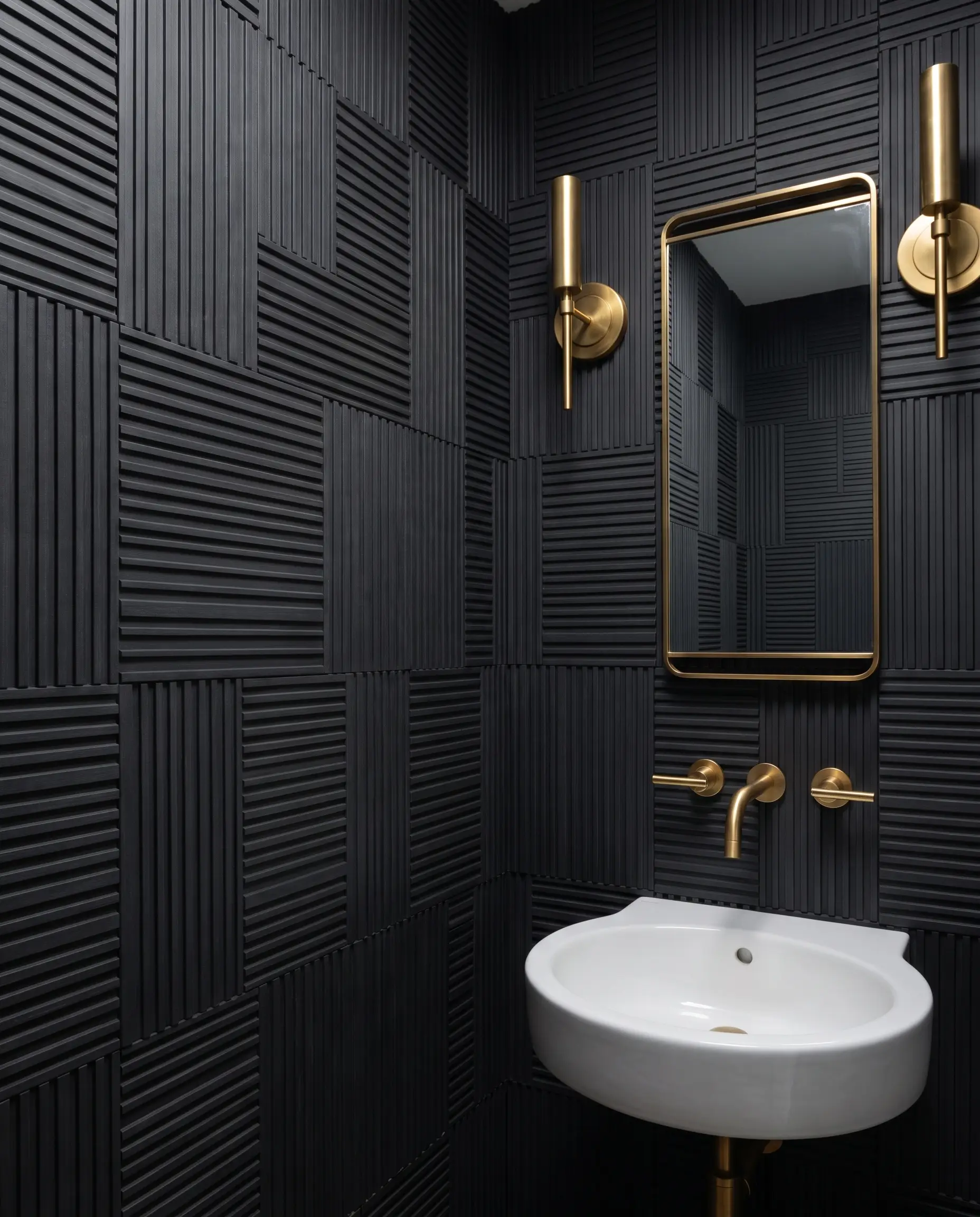 Deep matte charcoal waterproof WPC panels cover the walls of a small powder room featuring unlacquered brass wall-mounted faucets and a white ceramic sink.