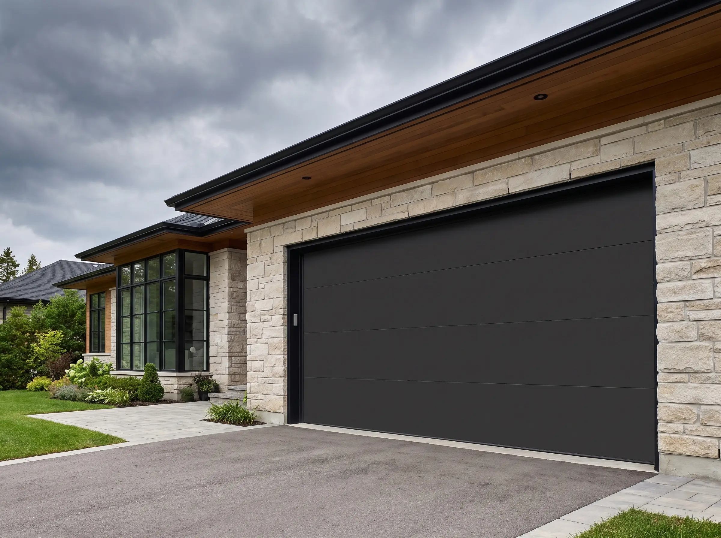 A flat flush-panel garage door painted in Colorbond Monument anchors a high-end exterior featuring natural light stone cladding, warm timber soffits, and black steel windows.