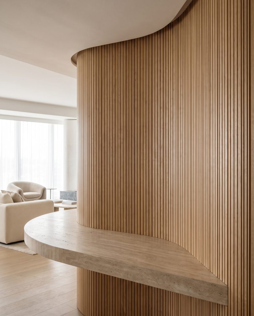 A sweeping curved architectural wall clad in flexible wooden wall panels anchors a thick honed travertine countertop acting as a sculptural entryway console.