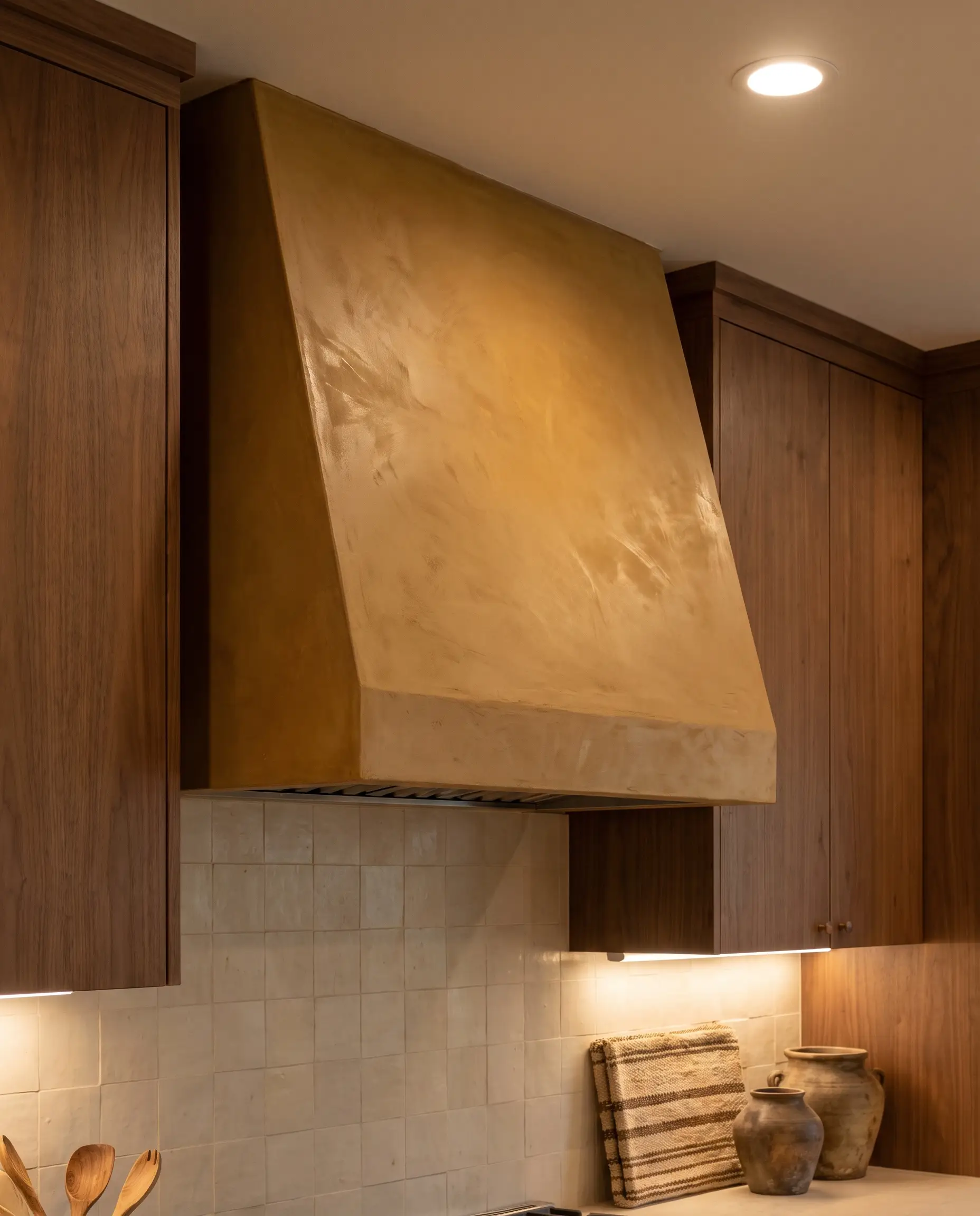 A sculptural sloped custom range hood finished in tinted earthy yellow lime plaster stands above a quiet neutral backsplash and warm wood cabinetry.