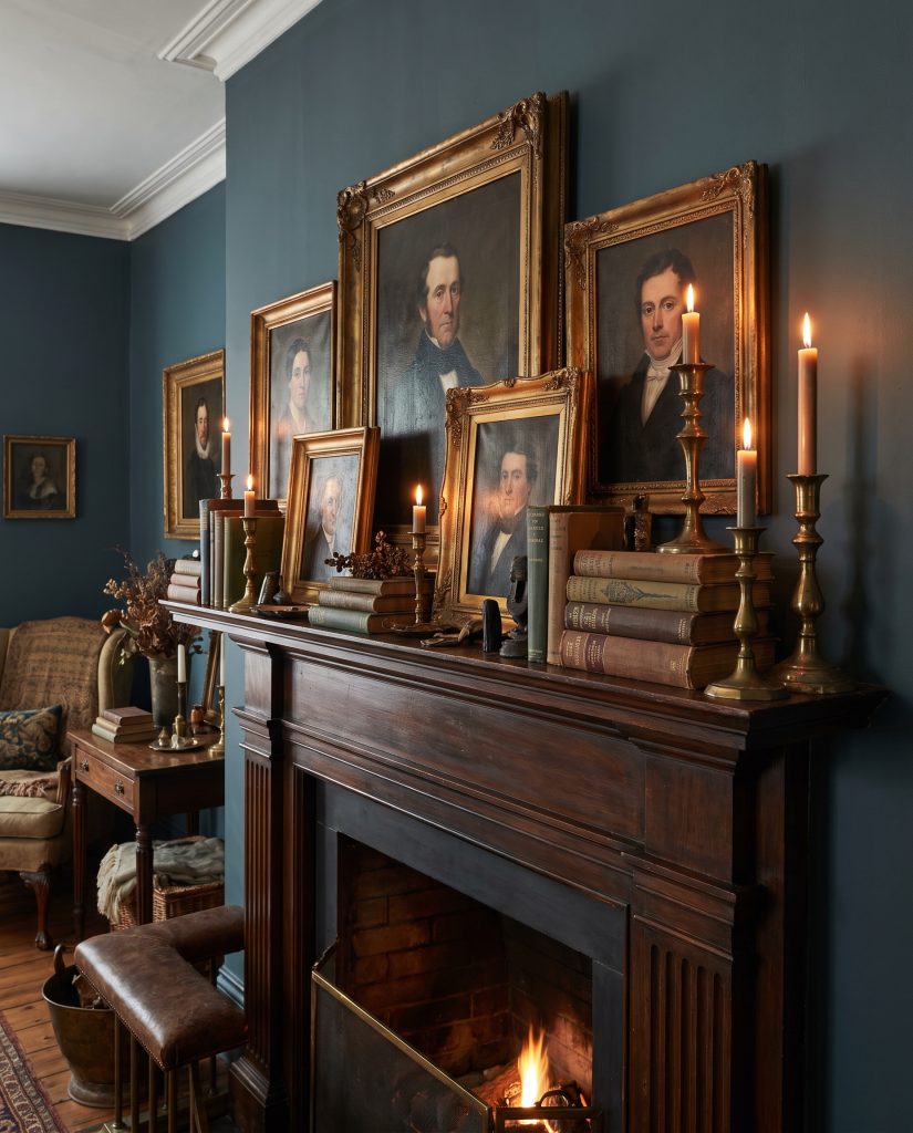 Antique oil portraits, oxidized brass candlesticks, and cloth-bound vintage books layer the ledge against a dramatic Farrow and Ball Hague Blue wall.