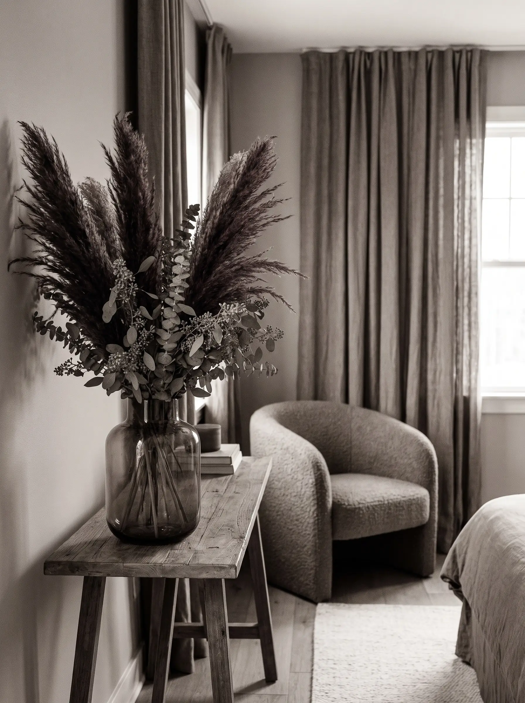 Dusty green seeded eucalyptus and dark plum pampas grass fill a smoked glass vase resting on a bedroom console near heavy drapes.