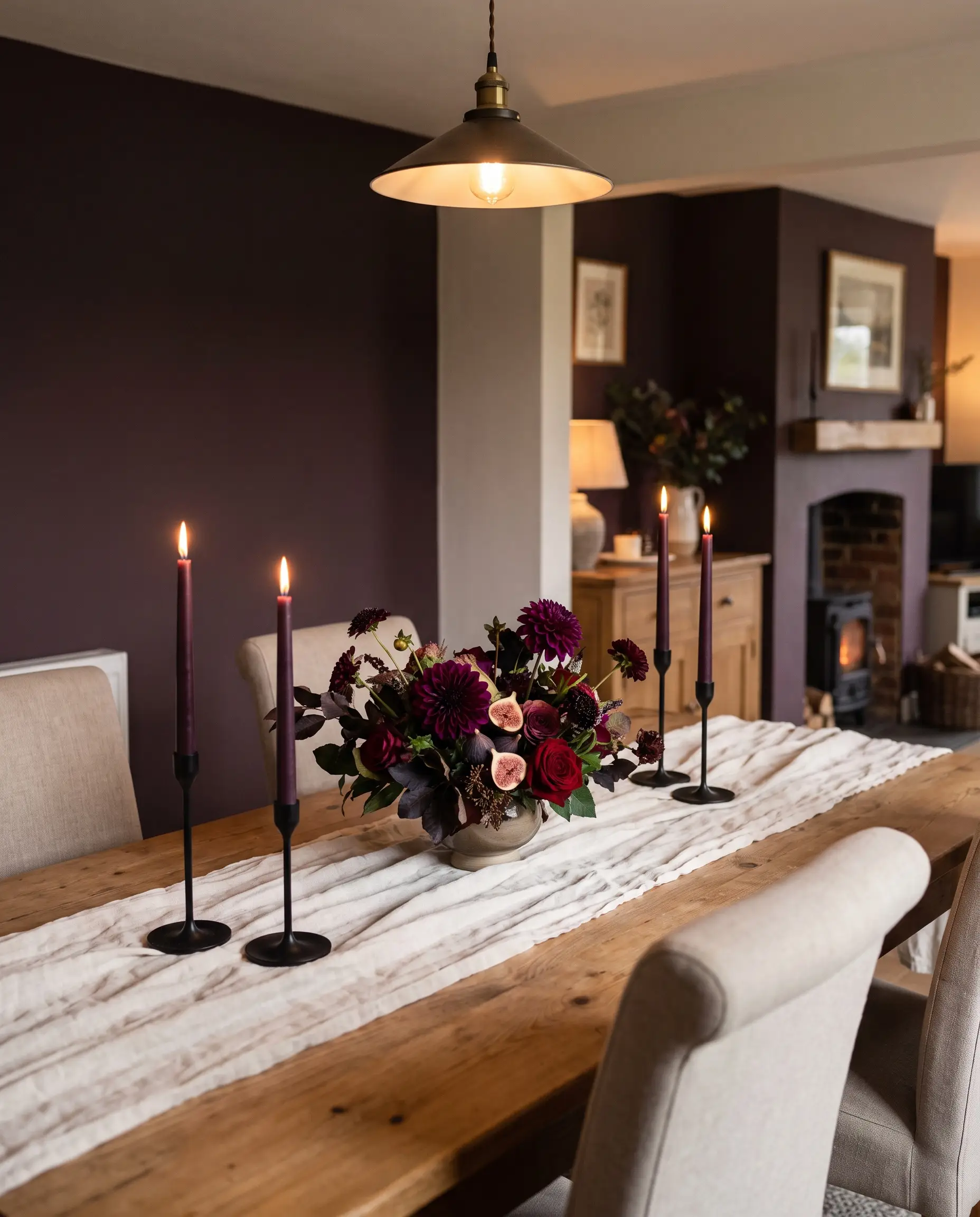 Deep aubergine dyed taper candles in iron holders stand on an ivory linen runner before a rich caponata painted accent wall and dark floral centerpiece.