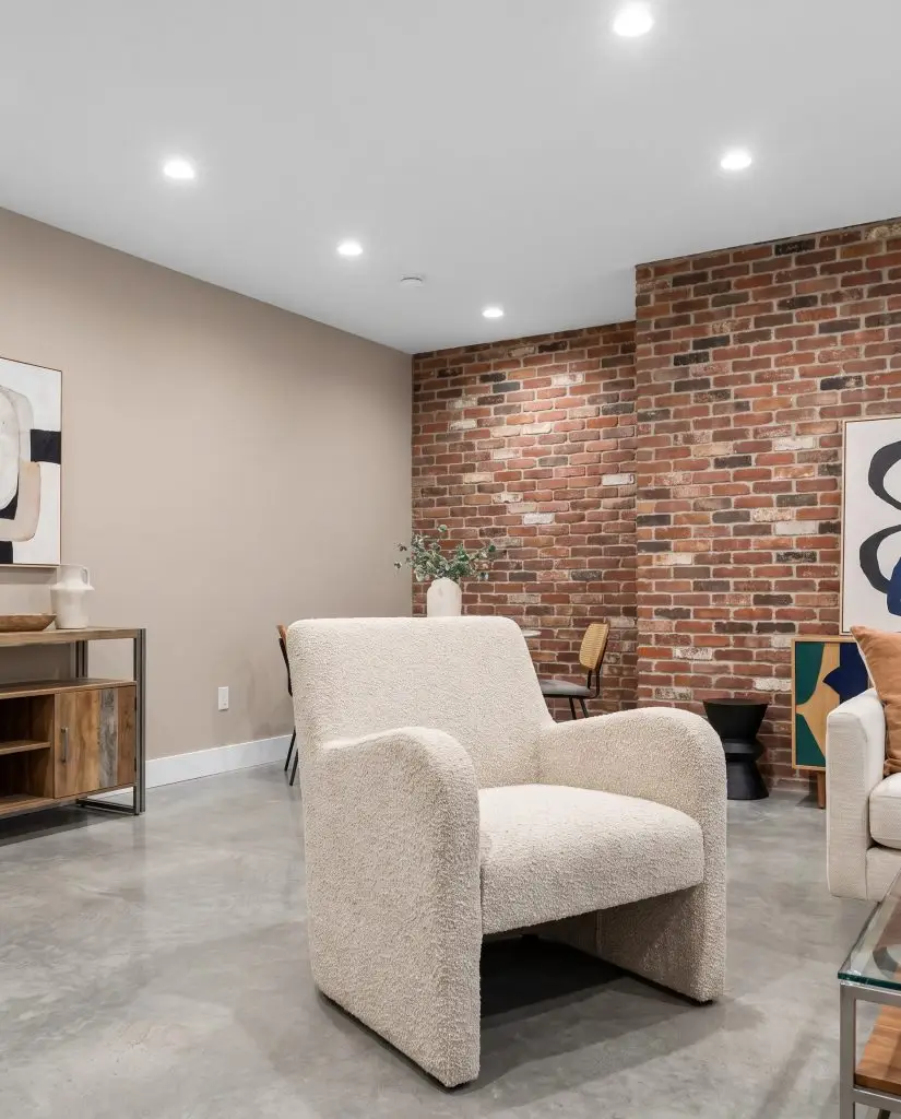 A bouclé fabric armchair sits on polished concrete flooring in a contemporary space featuring Farrow and Ball Elephant's Breath walls and reclaimed multi-colored red brick.
