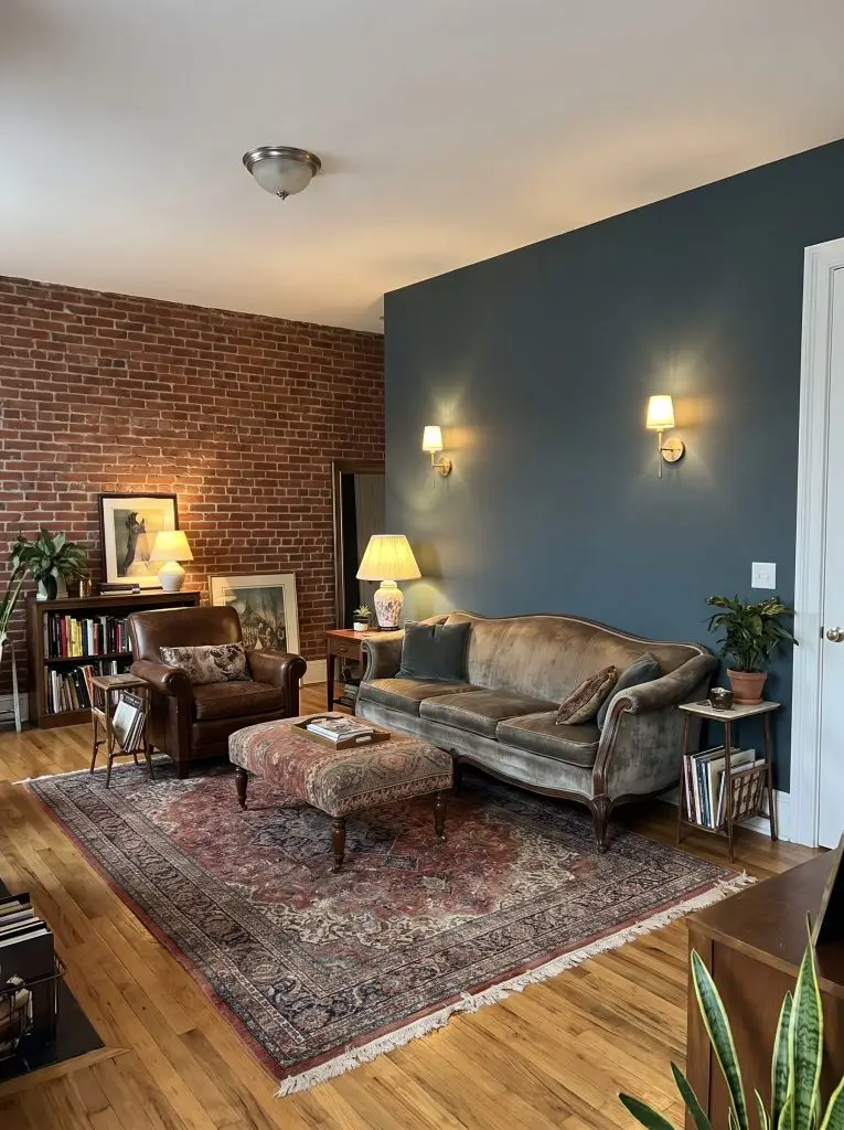 Unlacquered brass wall lamps cast a warm glow over a Persian rug, highlighting Farrow and Ball Hague Blue accent walls beside a red brick wall.