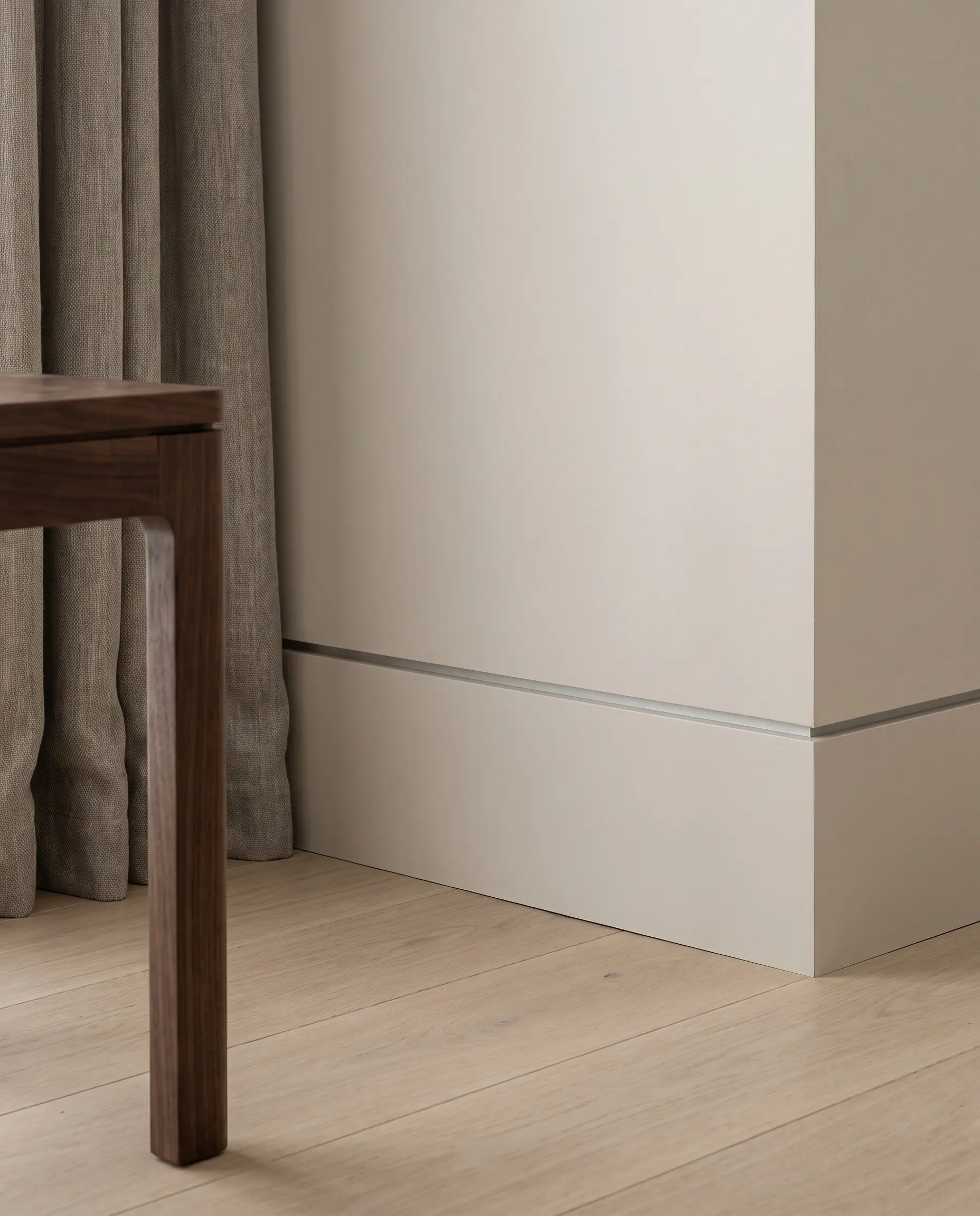 Flat-stock wood baseboards with an aluminum reveal bead create a subtle shadow gap above wide-plank oak flooring next to a solid walnut side table.