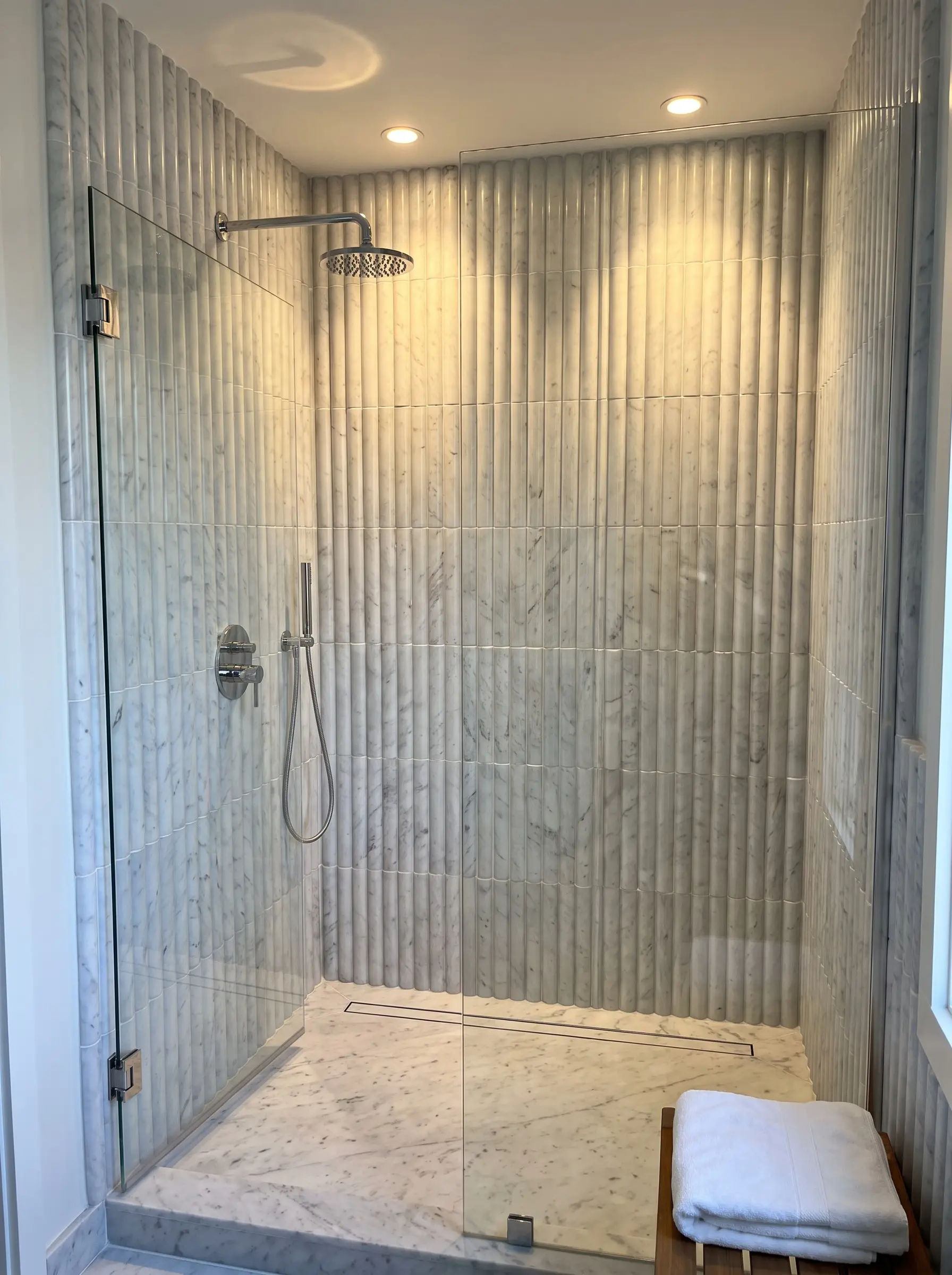 Vertical 3D fluted Carrara marble tiles set with bright white epoxy grout wrap a modern wet zone featuring polished chrome shower controls and a glass door.