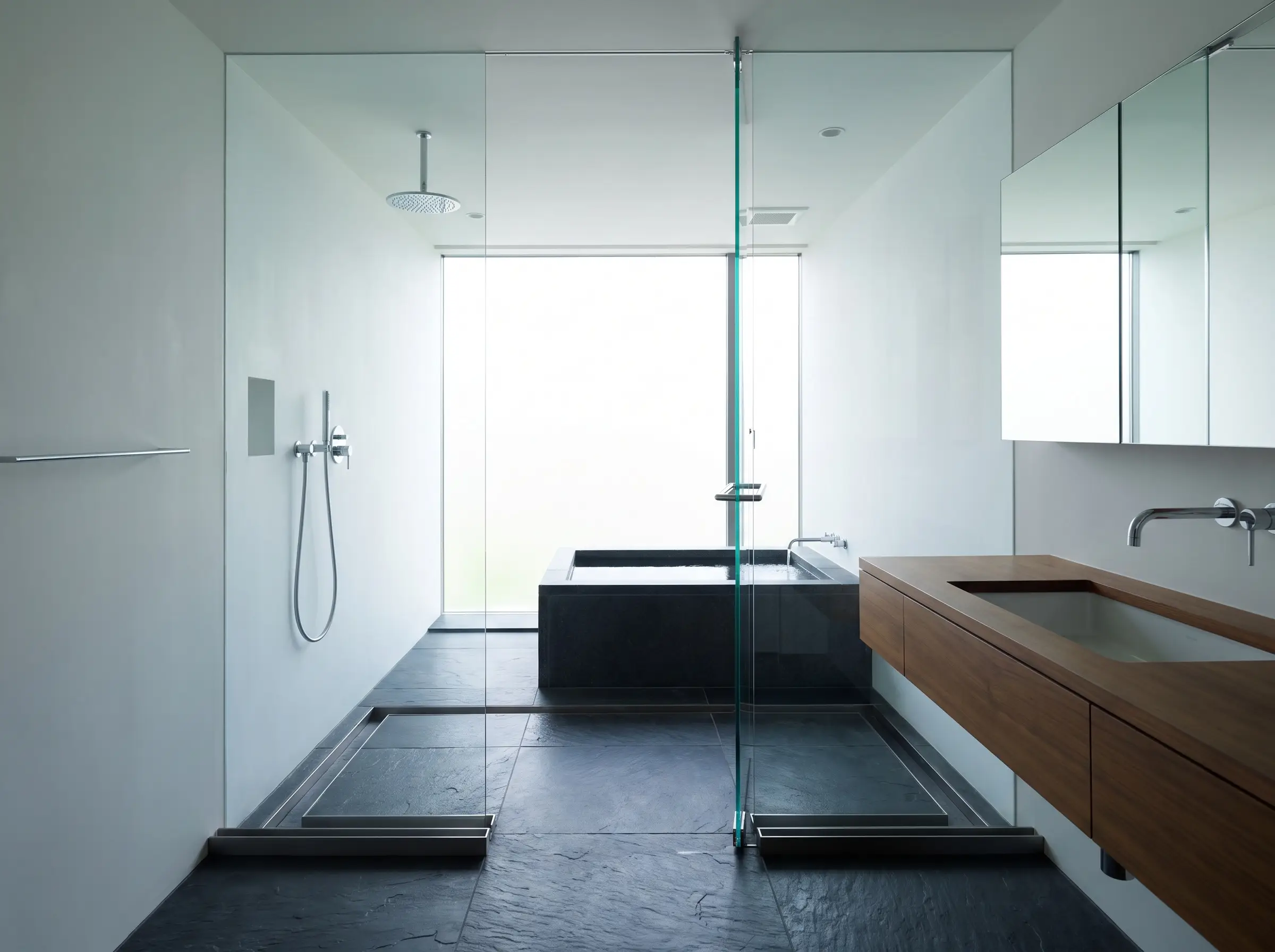 A floor-to-ceiling pane of thick tempered frameless glass separates a wet soaking zone from a dry vanity area over dark slate floor tiles.
