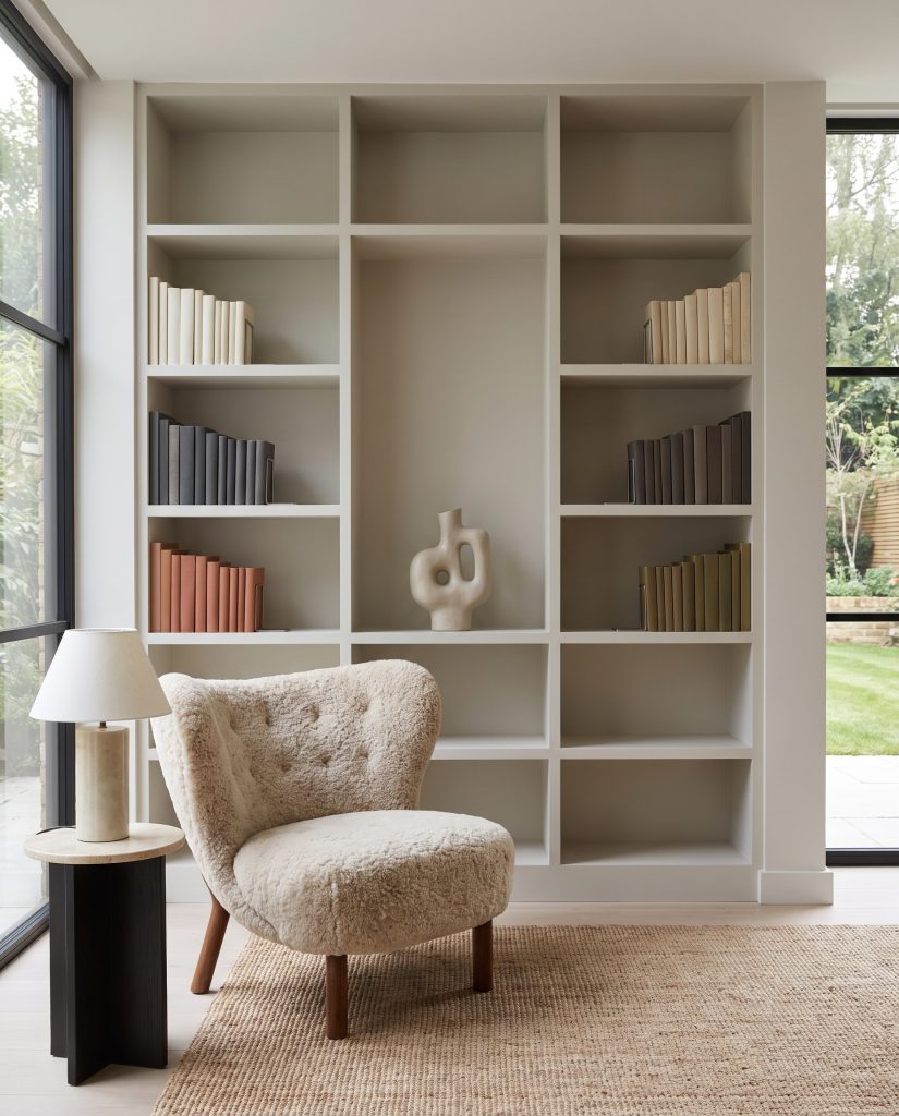 Built-in painted MDF open shelving displays color-blocked books and a single sculptural ceramic with generous negative space beside a cozy shearling chair.