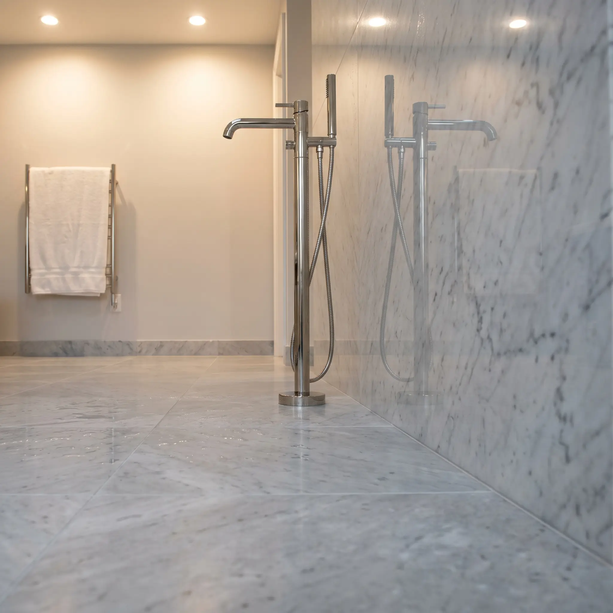 A velvety matte honed Carrara marble floor tile transitions seamlessly into a highly polished Carrara marble vertical wall slab beside a sleek polished nickel floor-mounted tub filler.