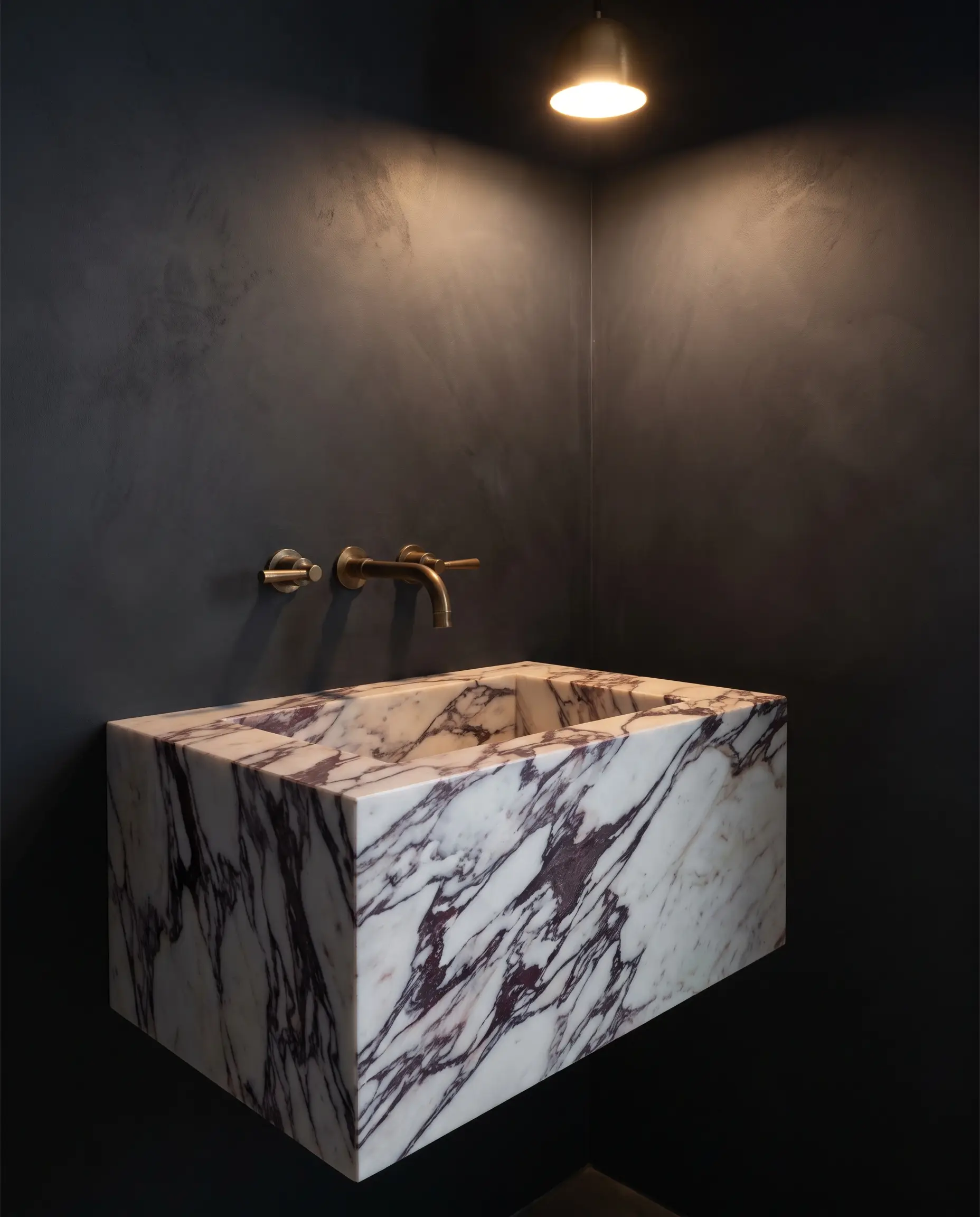 A sculptural Calacatta Viola marble plinth sink with heavy burgundy veining features unlacquered brass wall-mounted faucets against a dark charcoal wall in this marble bathroom design.