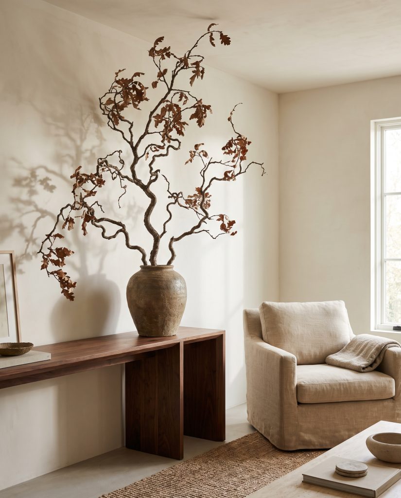 A single oversized foraged oak branch stands in a heavy raw stoneware vase on a solid walnut console table against a textured limewash wall.