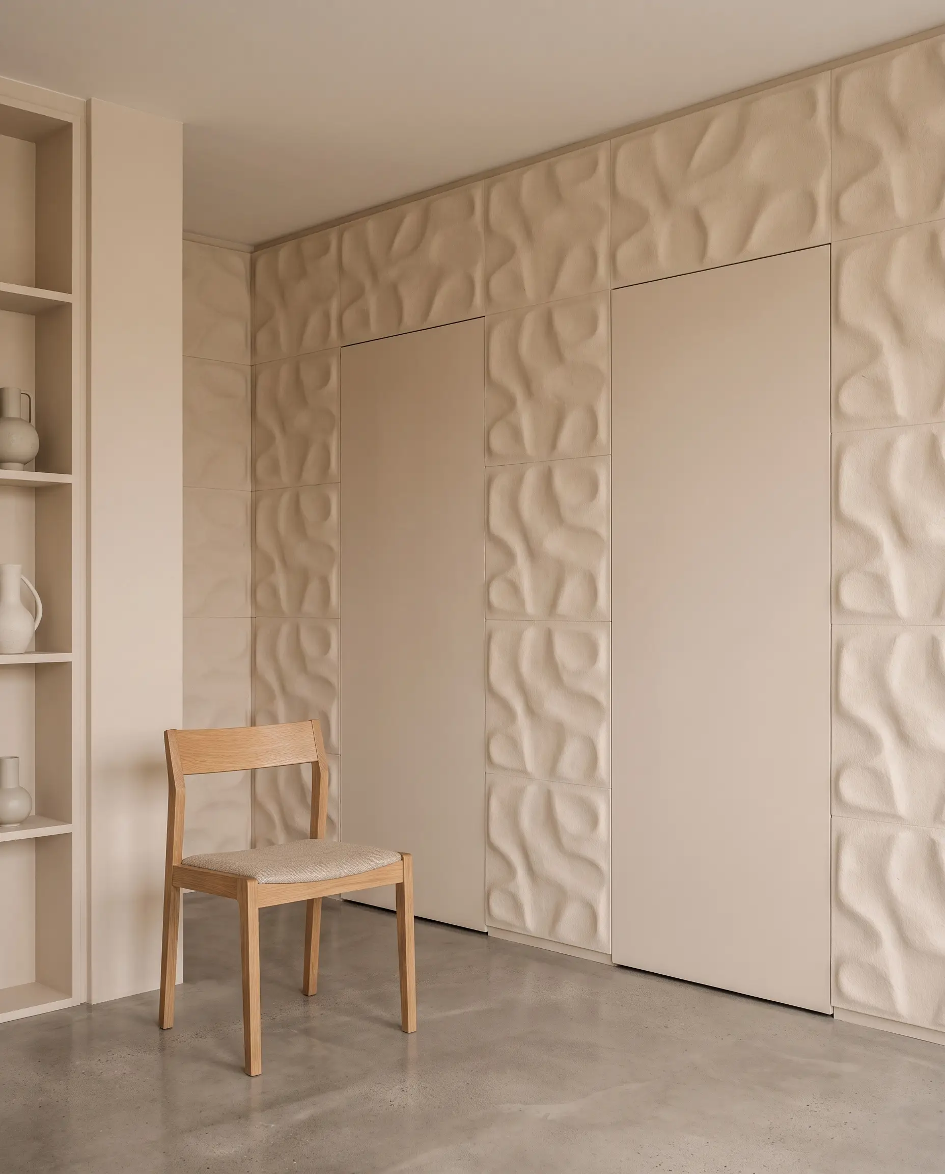 Flush-mount slab closet doors covered in seamless 3D wall panels blend into beige walls, creating a monolithic storage system in a minimalist bedroom.
