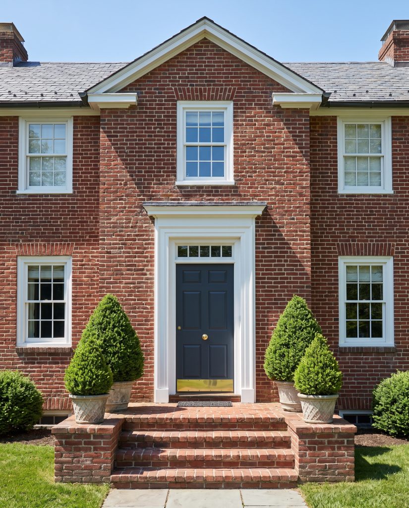 A highly polished unlacquered brass doorknob and kickplate highlight a deep Naval blue door set against glossy white trim and a red brick facade.