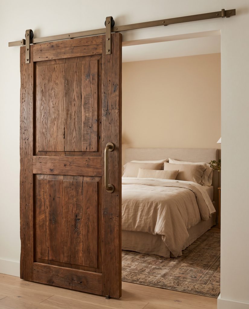 A heavy antique reclaimed wood sliding barn door with matte bronze hardware opens into a cozy modern farmhouse bedroom featuring a linen-draped bed.