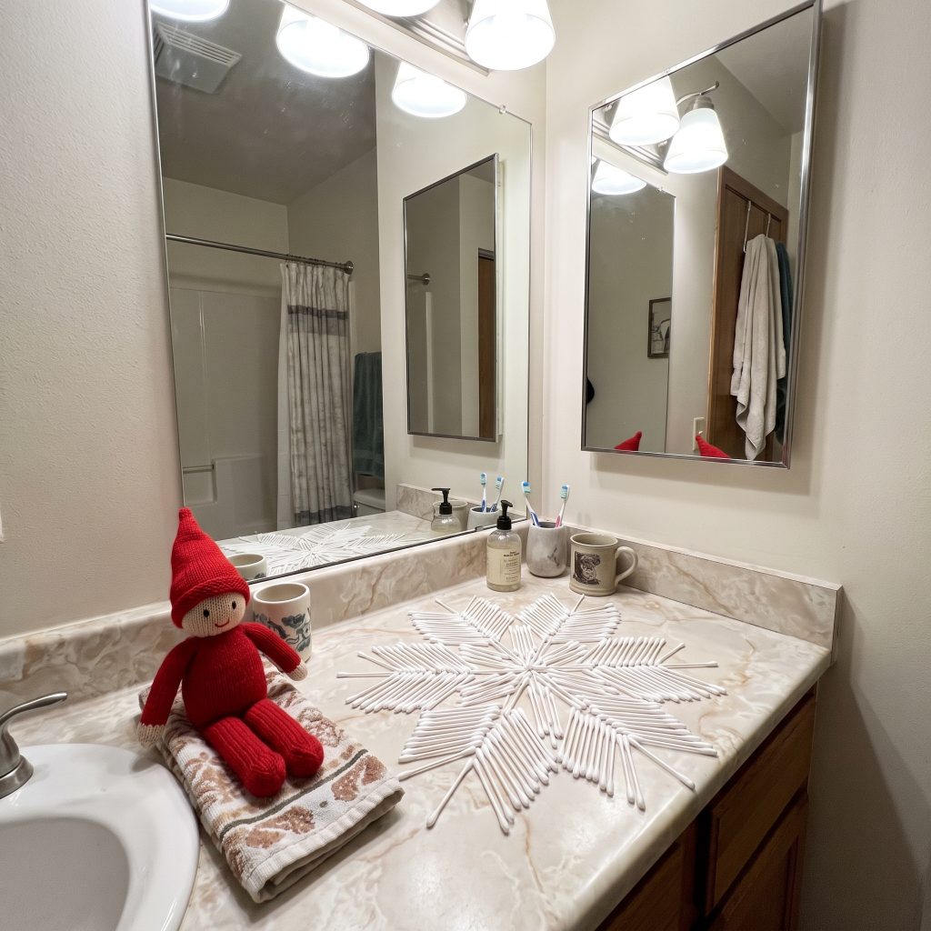 A red holiday Scout Elf sits next to a geometric snowflake pattern meticulously arranged out of white cotton swabs on a standard bathroom counter.