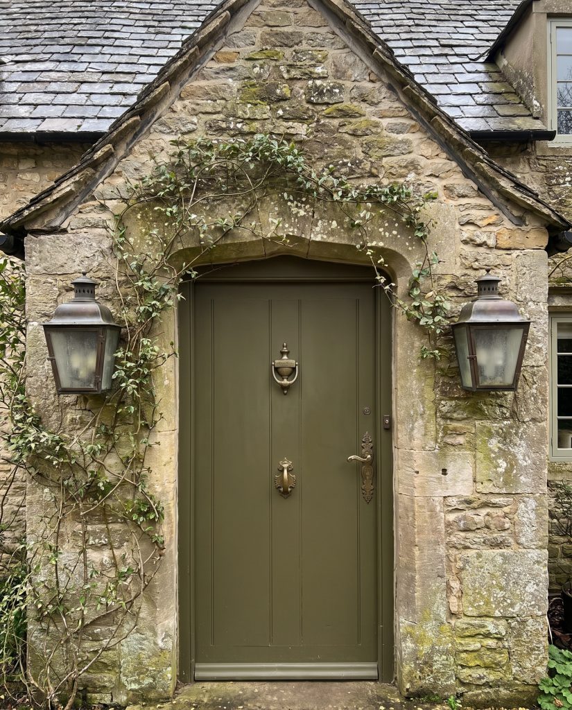 Climbing ivy creeps up a rough-cut natural stone facade framing a Secret Garden green door with heavy aged brass hardware and bronze sconces.