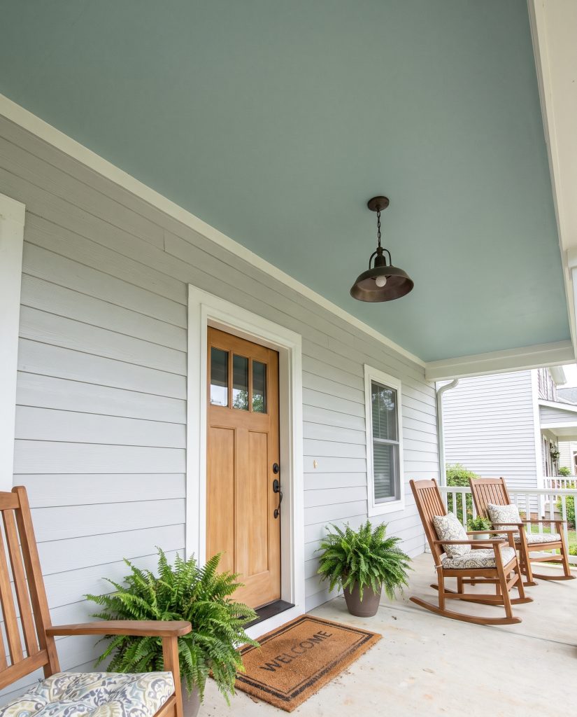 A serene blue-green painted porch ceiling draws the eye upward above modest gray siding and a simple wooden front door in a covered entryway.