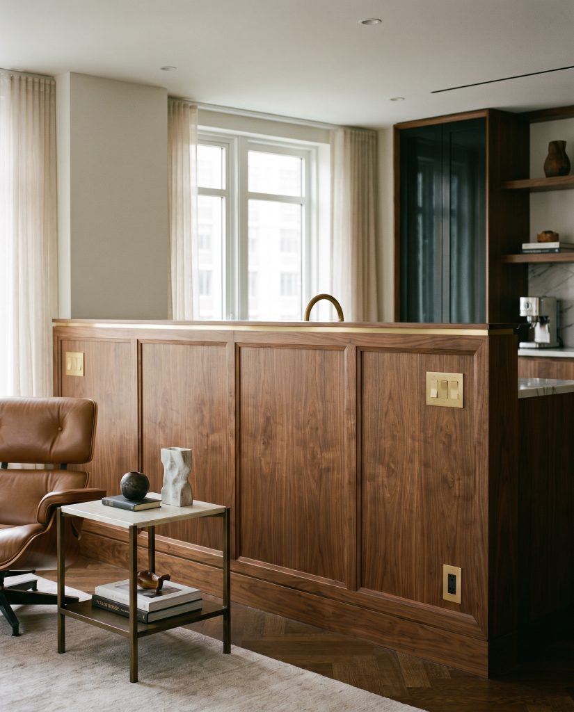A flawless half-wall of solid walnut paneling features a custom milled brass U-channel trim rail echoing integrated brass switch plates beside a refined lounge chair.