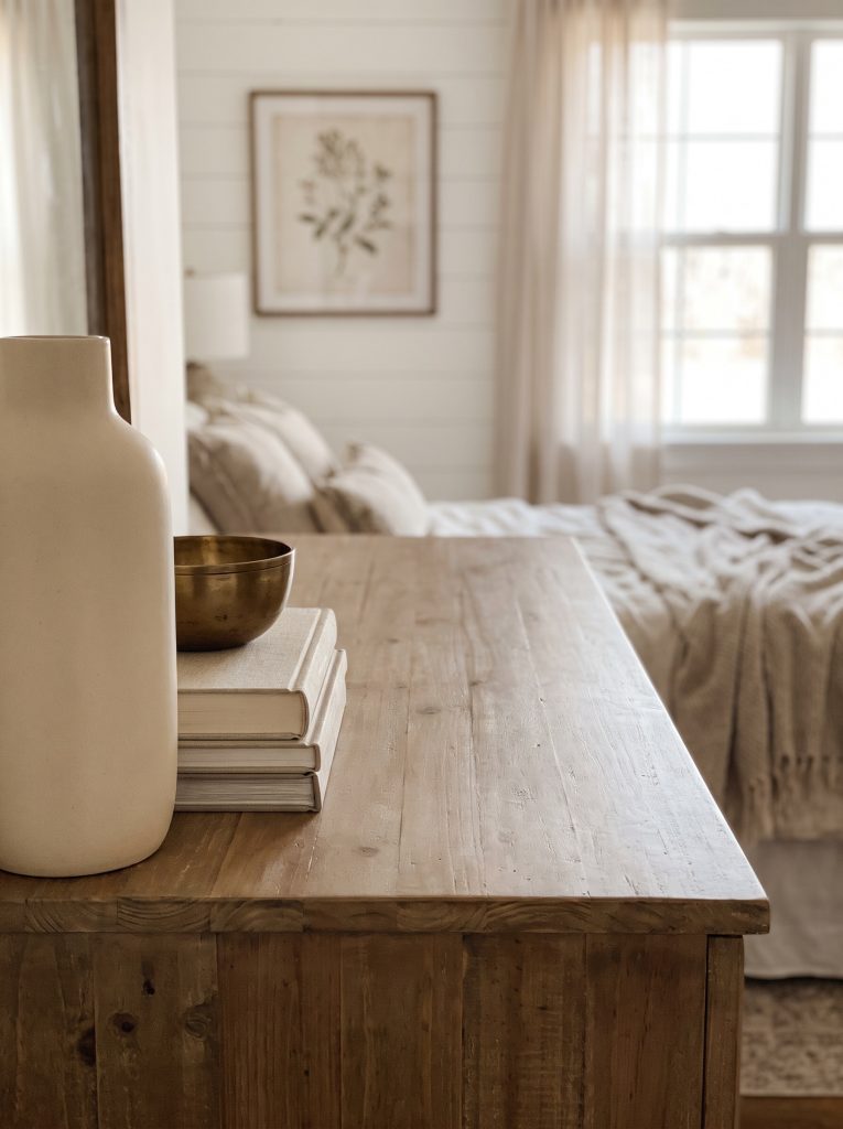 A wooden bedroom dresser displays a tall matte ceramic vase, linen-bound books, and a low brass bowl grouped with intentional negative space.