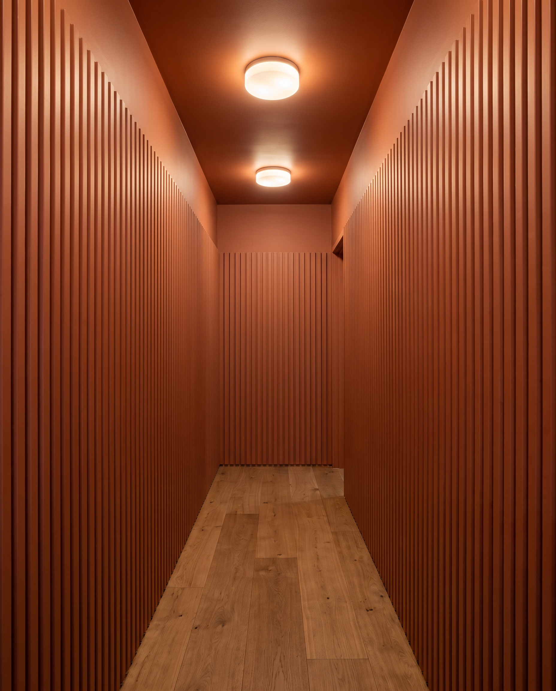 Deeply grooved vertical fluted 3D wall panels color-drenched in warm terracotta line a narrow hallway with minimalist oak flooring and flush-mount lighting.
